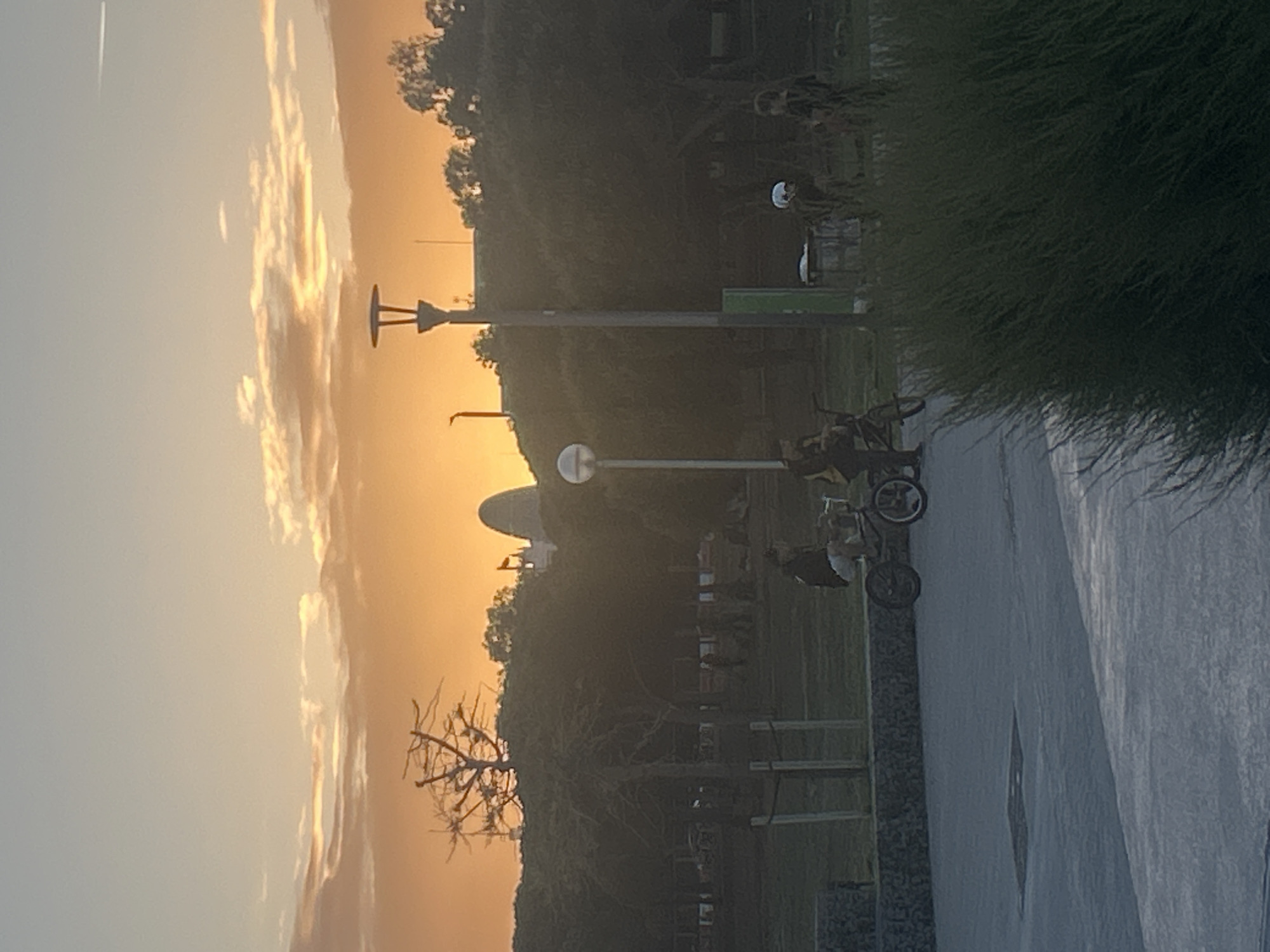 The image shows a sunset scene in Barcelona, Spain. A paved path runs through a park, with trees lining the edge. Lampposts stand along the path, and several bicycles are parked nearby. The sky is filled with orange and yellow hues from the setting sun. In the background, a building with a dome can be seen.