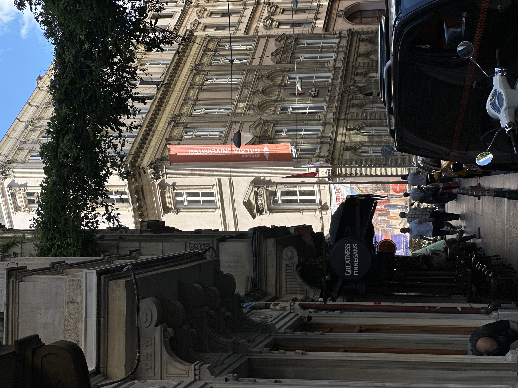 The image captures a street scene in Beyoğlu, Turkey. In the foreground, the facade of a building with ornate architectural details, including decorative carvings and columns, is visible on the left. A dark awning extends over a sidewalk, beneath which is a sign reading "GREATNESS FINNEGAN'S". Several people are walking on the sidewalk, and a person is partially visible in a bent-over posture on the far left. Further down the street, another building with multiple windows and architectural embellishments is prominent, with an orange banner hanging vertically that reads "GALATA ÜNİVERSİTESİ" (Galata University). The street is lined with more buildings, some with visible window frames and classical architectural elements. Vehicles, including a black van and a white scooter, are parked along the curb. Trees with dark green foliage are seen overhead, casting shadows. The sky is a clear blue with scattered white clouds. The overall impression is that of a bustling urban environment with historical architecture.