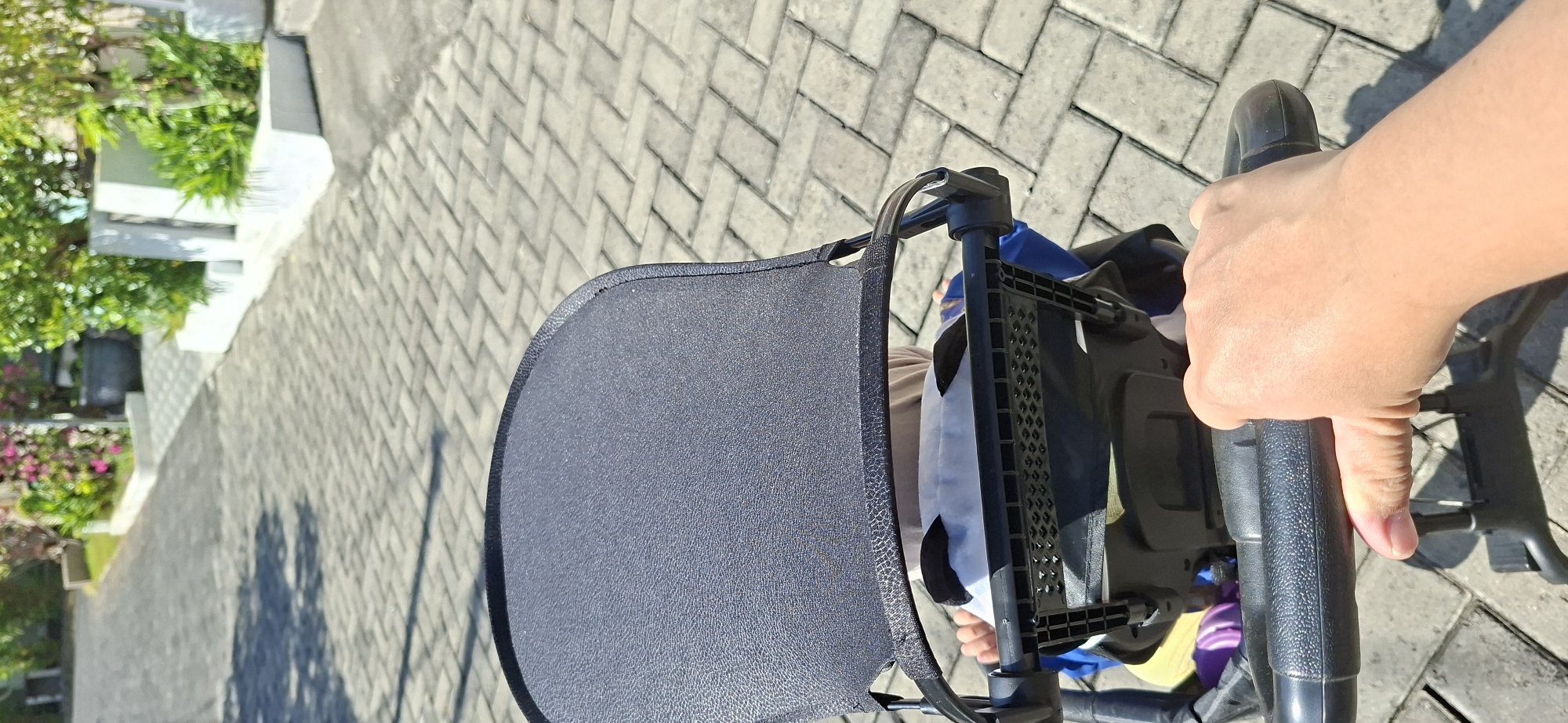 A close-up POV shot shows a person's hand gripping the handle of a baby stroller on a sunny day. The stroller's dark grey canopy is prominently featured in the foreground, with a glimpse of a baby inside wearing a panda-themed pillow. The stroller's frame and handle are made of black plastic. The background consists of a grey brick-paved pathway with dappled sunlight and shadows. Beyond the pathway, there is a raised garden bed with lush green plants and some pink flowers, suggesting a residential or park area in Surabaya, Indonesia. The overall scene is peaceful and suggests an outing.