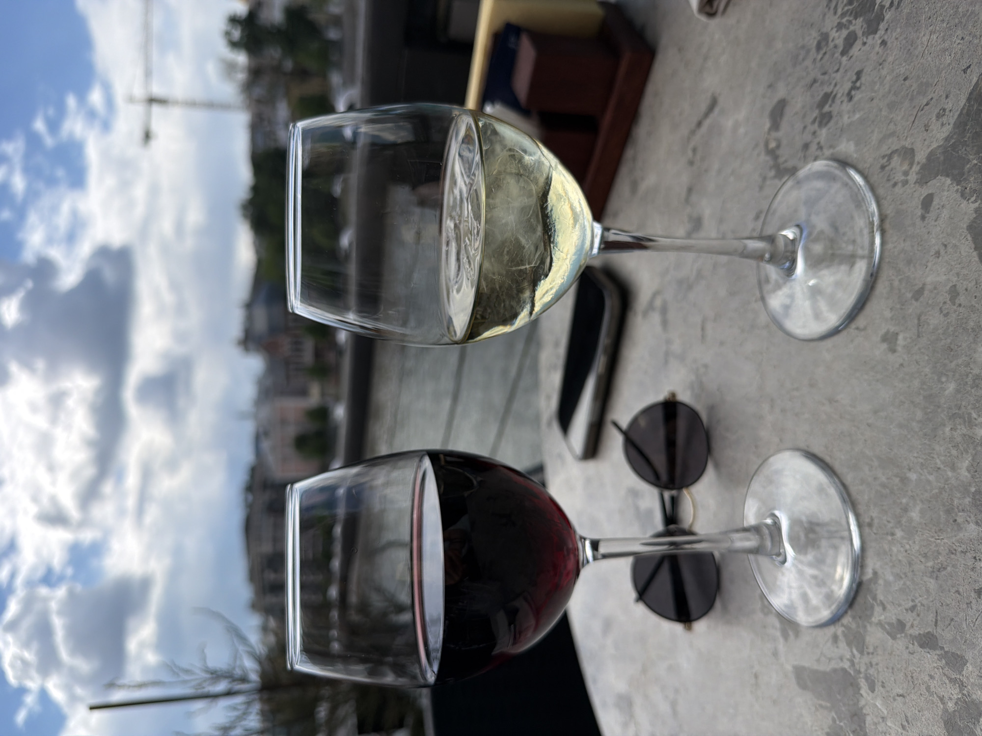 The image depicts two wine glasses, one filled with red wine and the other with white wine, resting on a grey, textured surface, likely a table. Between the glasses lie a pair of sunglasses and a smartphone. The backdrop reveals a cityscape under a partly cloudy sky, including a crane. The location is the city of Unknown, Unknown.