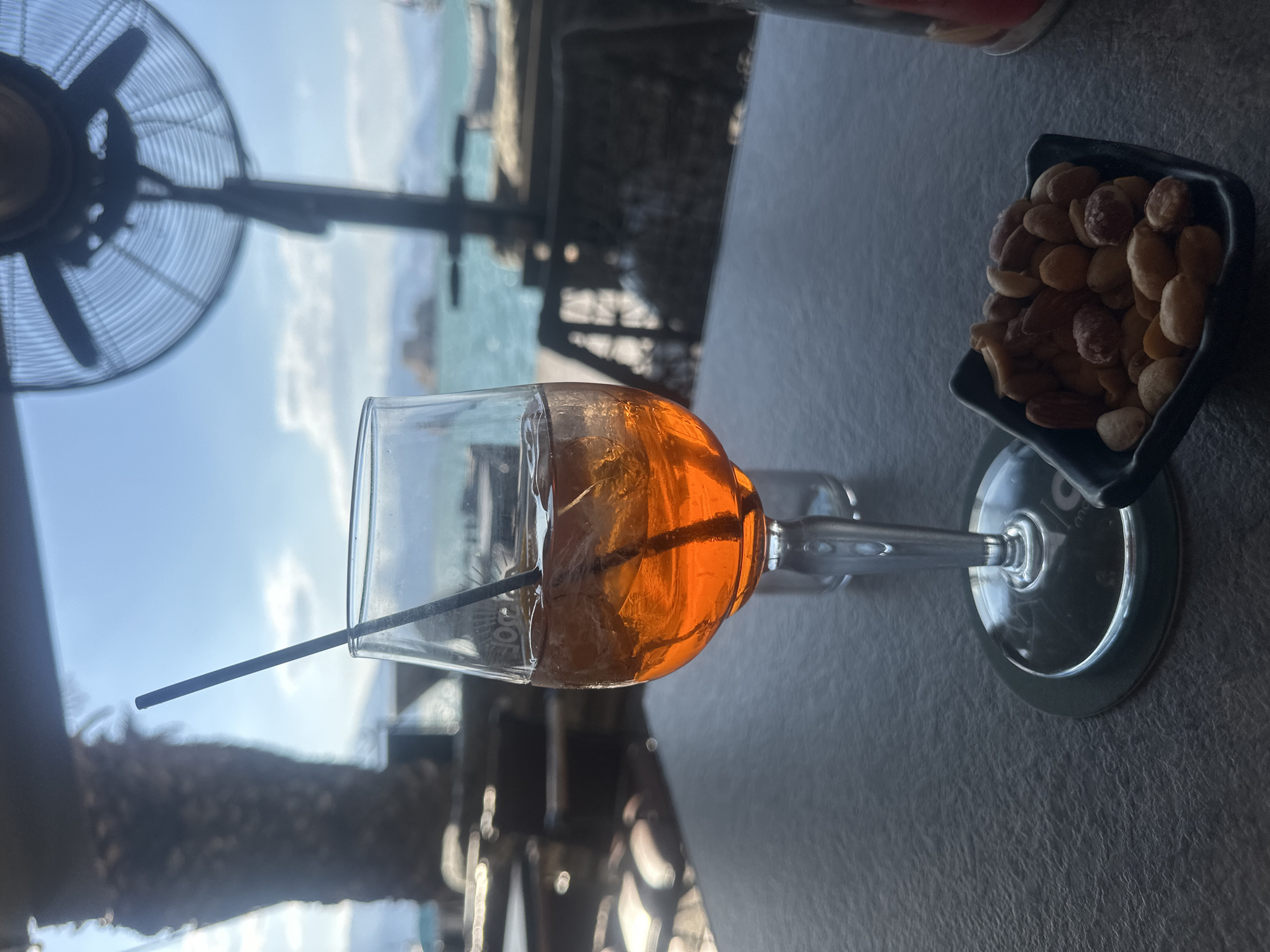 A glass of an orange aperitif with ice and a straw sits on a dark table, with a small dish of mixed nuts to its right. In the background, the turquoise sea is visible, with a distant fortress on a rocky outcrop. A large oscillating fan is positioned in the upper right, and a blurry palm tree is in the upper left. The scene appears to be an outdoor seating area of a restaurant or bar, likely in Nafplio, Greece, given the presence of the fortress. The lighting suggests it is daytime.