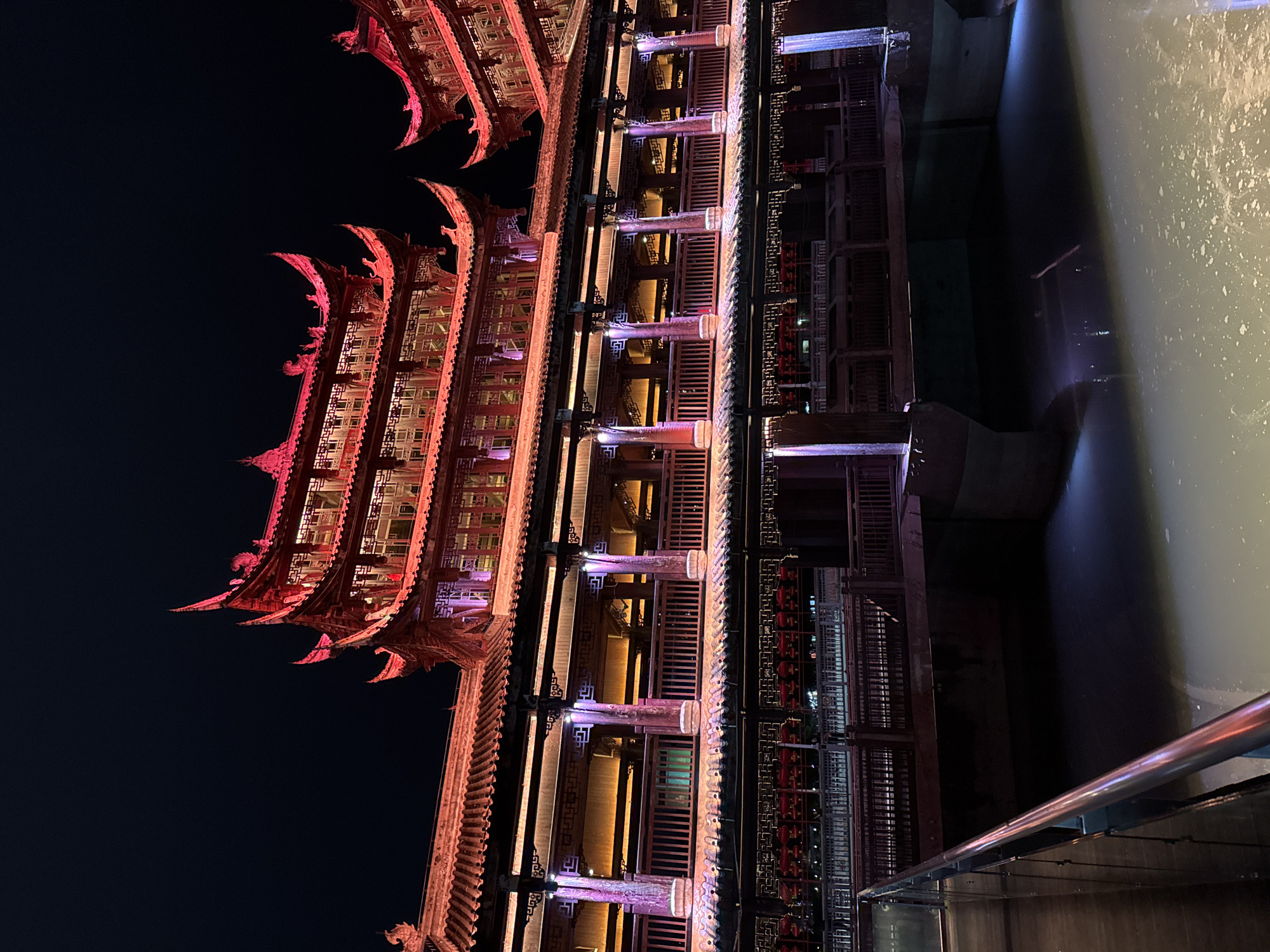 The image depicts a multi-tiered Chinese building illuminated at night, likely located in Qianjiang, China. The building features traditional architectural elements such as ornate eaves, wooden structures, and decorative details. It is lit up with red, orange, and purple lights, creating a vibrant and colorful display against the dark night sky. The building is situated alongside a body of water. A metal railing is visible in the foreground on the right, suggesting the photo was taken from a boat or a platform along the water's edge. The water reflects some of the building's lights.