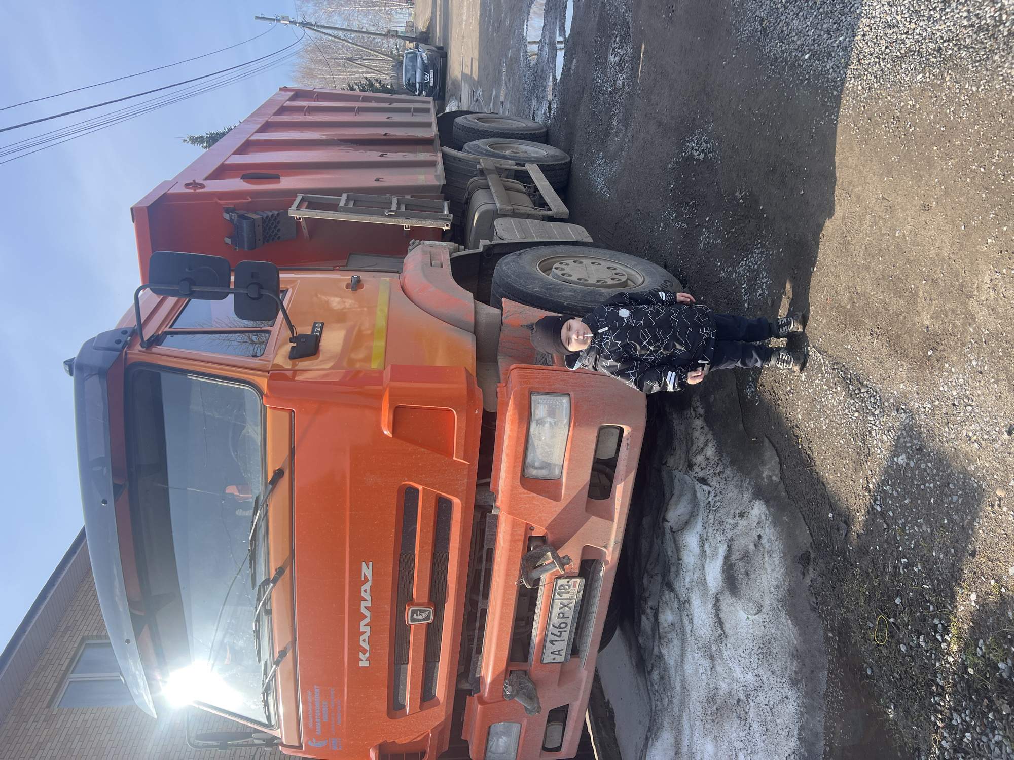 A boy stands next to an orange Kamaz dump truck in Chaikovsky, Russia. The truck's license plate reads "A 146 PX 18". The location appears to be a street with a mixture of asphalt and dirt, with some melting snow present. The sky is clear and blue. A residential building is partially visible in the lower left corner. Another vehicle is parked further down the street.