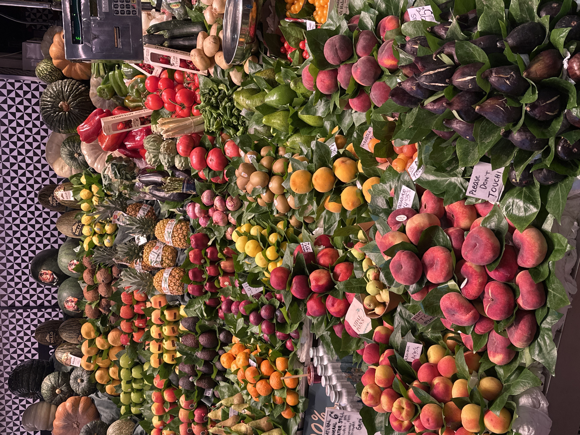 This is a close-up, eye-level, full-frame photograph of a vibrant fruit and vegetable market stall, likely in Barcelona, Spain, given the signage that includes Spanish text. The foreground is dominated by a profusion of fresh produce artfully arranged on display.
In the lower right and center, there are piles of dark purple figs, their skins marked with lighter lines. To their left and above, a large quantity of flat, reddish-purple peaches (often called Saturn peaches or doughnut peaches) are mounded on green leaves. Further to the left, there are many round, red peaches and nectarines. Above these, stacks of bright orange tangerines and some lighter orange apricots are visible, also arranged on green leaves. Scattered throughout the display are pineapples, pears, apples, kiwis, and various colorful peppers.
In the background, the stall displays larger items such as pumpkins, melons, and gourds. Some of the melons have labels with Spanish text, one reading "BOLLO". There are also crates of tomatoes and other vegetables. A portion of a digital scale with a display showing "PRECIO kg" (price per kg) is visible on the far right, suggesting a commercial transaction is taking place or has taken place. The backdrop of the stall features a repeating geometric pattern of black and white squares, typical of market aesthetics. The lighting is bright, highlighting the rich colors and textures of the produce. No people are clearly visible in the frame.