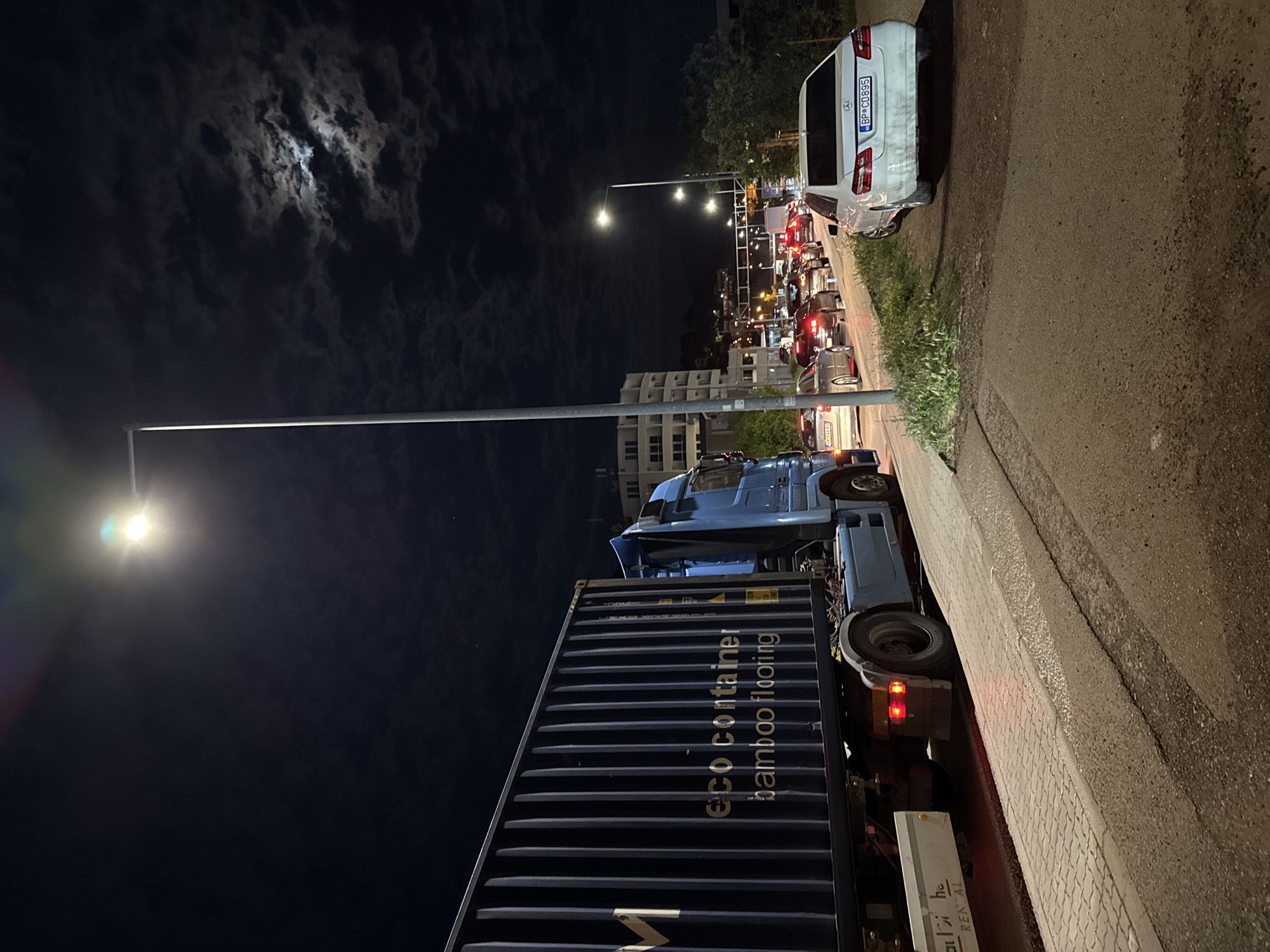 This image captures a nighttime street scene in Zankovići, Montenegro, characterized by a line of traffic and artificial illumination. In the foreground, a large blue semi-truck with a dark blue shipping container is visible on the right side of the road. The container prominently displays the text "eco container bamboo flooring." Further down the road, a white Mercedes-Benz minivan/van is parked or stopped on the right, its license plate "BP C0895" clearly legible.
A queue of vehicles with their red taillights glowing stretches into the distance, suggesting heavy traffic or a standstill. The scene is lit by several streetlights on tall poles, casting bright light onto the road and the side of the truck. In the background, residential buildings can be vaguely seen under the dim light. The sky is dark but features visible clouds, with a bright moon shining through them. The ground consists of asphalt road, a paved sidewalk, and some patches of grass. The overall atmosphere is one of a busy street at night.