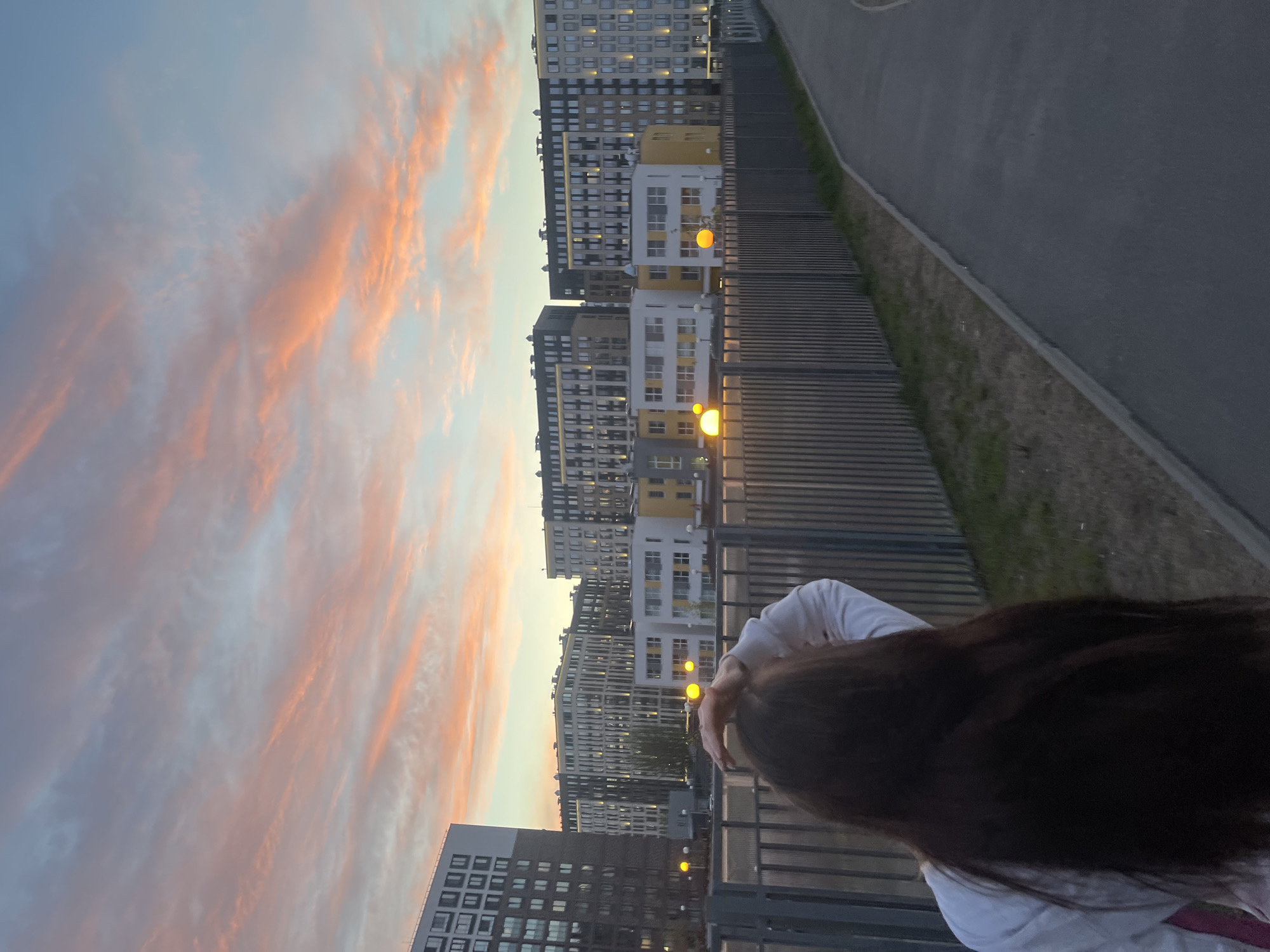 The image shows a woman with long, dark hair standing in front of a metal fence, with a street and tall apartment buildings in the background. The sky is visible above the buildings, displaying a sunset with pink and orange clouds. Street lights are illuminated in front of some of the apartment buildings. The location is likely Saint Petersburg, Russia, based on the architecture of the buildings.