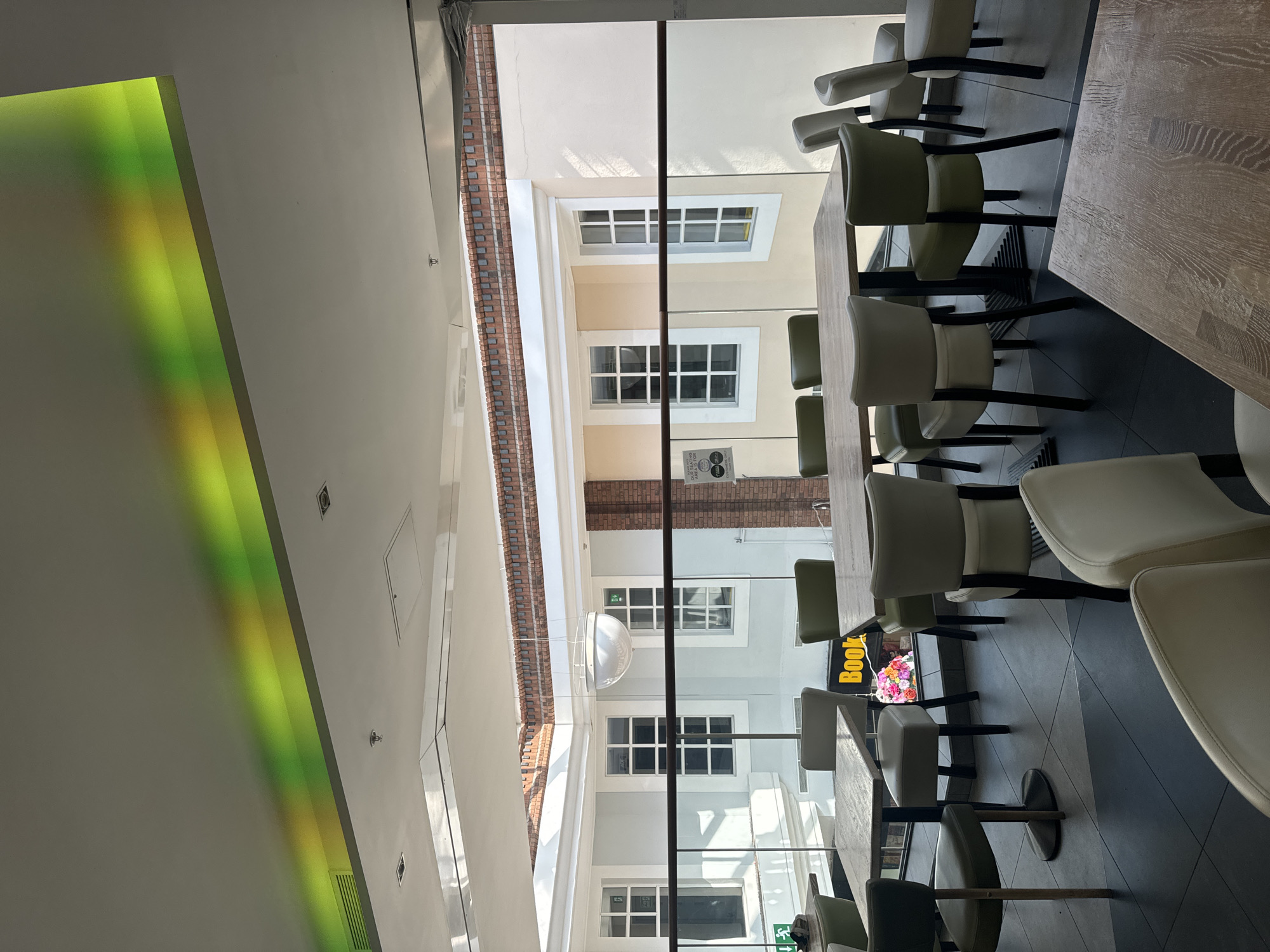 The image depicts an indoor seating area, likely a cafe or restaurant, with a view into an internal courtyard. The foreground features several tables and chairs. The tables are made of wood, with some having dark metal bases. The chairs are upholstered in neutral tones of beige and green.
Beyond the seating area, a glass partition separates the indoor space from an internal courtyard. The courtyard is characterized by light-colored walls, multiple windows with white frames, and a brick facade at the top. A large, spherical white light fixture hangs from the ceiling in the courtyard. Above the courtyard and extending into the foreground, there is a translucent ceiling element with colored lighting, casting green and yellow hues. A sign that appears to read "Book" is visible in the background, suggesting a bookstore or a section within a establishment.
The overall impression is one of a modern, well-lit interior space designed for dining or socializing, with a connection to an architectural courtyard. The location in the city of Waterford, Ireland, has not been confirmed by any visual elements within the image.