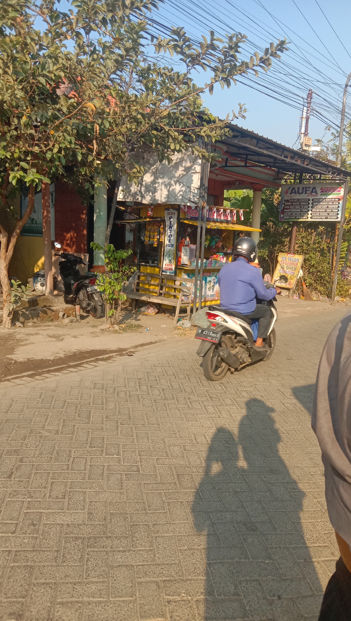 In Curah Dukuh, Indonesia, a man wearing a blue long-sleeved shirt and a helmet rides a white motorcycle down a brick-paved street. The motorcycle is heading away from the camera. In the background, a small open-air shop with a yellow awning sells various goods, including food items and possibly printing services, indicated by signs. A black motorcycle is parked to the left of the shop. The scene is set under a clear blue sky with sunlight casting long shadows on the street, suggesting it is late afternoon. A tree with green leaves shades part of the shop and the street. The overall environment appears to be a local street or market area.