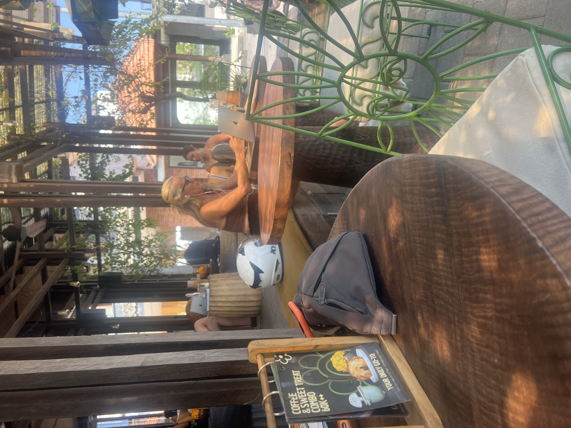 A woman with blonde hair and glasses, wearing a brown tank top, sits at a wooden table outdoors in a cafe. She is looking at a laptop and appears to be typing. In front of her is a laptop, a water bottle, and a white motorcycle helmet on a yellow cushion. A dark gray fanny pack with an orange strap lies on the table to her left. Also on the table is a wooden stand holding a menu that reads "COFFEE & SWEET TREAT COMBO 60K++ YOUR DAILY GO-TO". Behind her, another woman with dark hair is seated at a different table, partially visible. The cafe appears to be open-air with a wooden roof structure and greenery. The setting suggests a relaxed atmosphere, possibly a tourist area, given the helmet. The city of Mengwi, Indonesia is known for its cultural sites and temples, but this scene depicts a more modern cafe setting within what could be a commercial or resort area. The lighting indicates it is daytime, with sunlight filtering through the canopy.