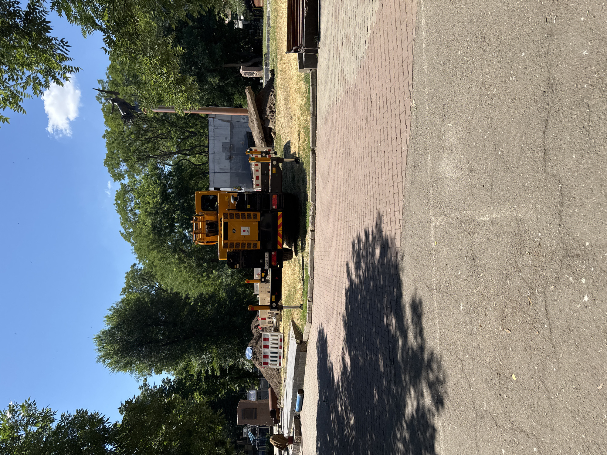 The image shows a sunny day with a clear blue sky and scattered white clouds. The scene appears to be an outdoor area, possibly a park or a street undergoing construction, in Odesa, Ukraine.
In the foreground, a paved area transitions into a textured grey asphalt surface. The paved area is made of reddish-brown interlocking bricks, casting dappled shadows of tree leaves.
Further back, on the left side of the image, a yellow truck with construction equipment is visible. It appears to be a crane truck with an extended arm. The truck has some signage with text, including "42002 BH". Next to the truck, there's a pile of dirt, and a section of red and white striped barrier, indicating ongoing work or repair. A large tree trunk lies on the ground near the construction area.
Beyond the construction site, lush green trees with dense foliage dominate the background, suggesting a park or a tree-lined street. The sun is high in the sky, casting distinct shadows, implying it is likely midday. The overall impression is one of an active construction project taking place in a green, urban environment on a bright, clear day. No people are clearly visible in the image.