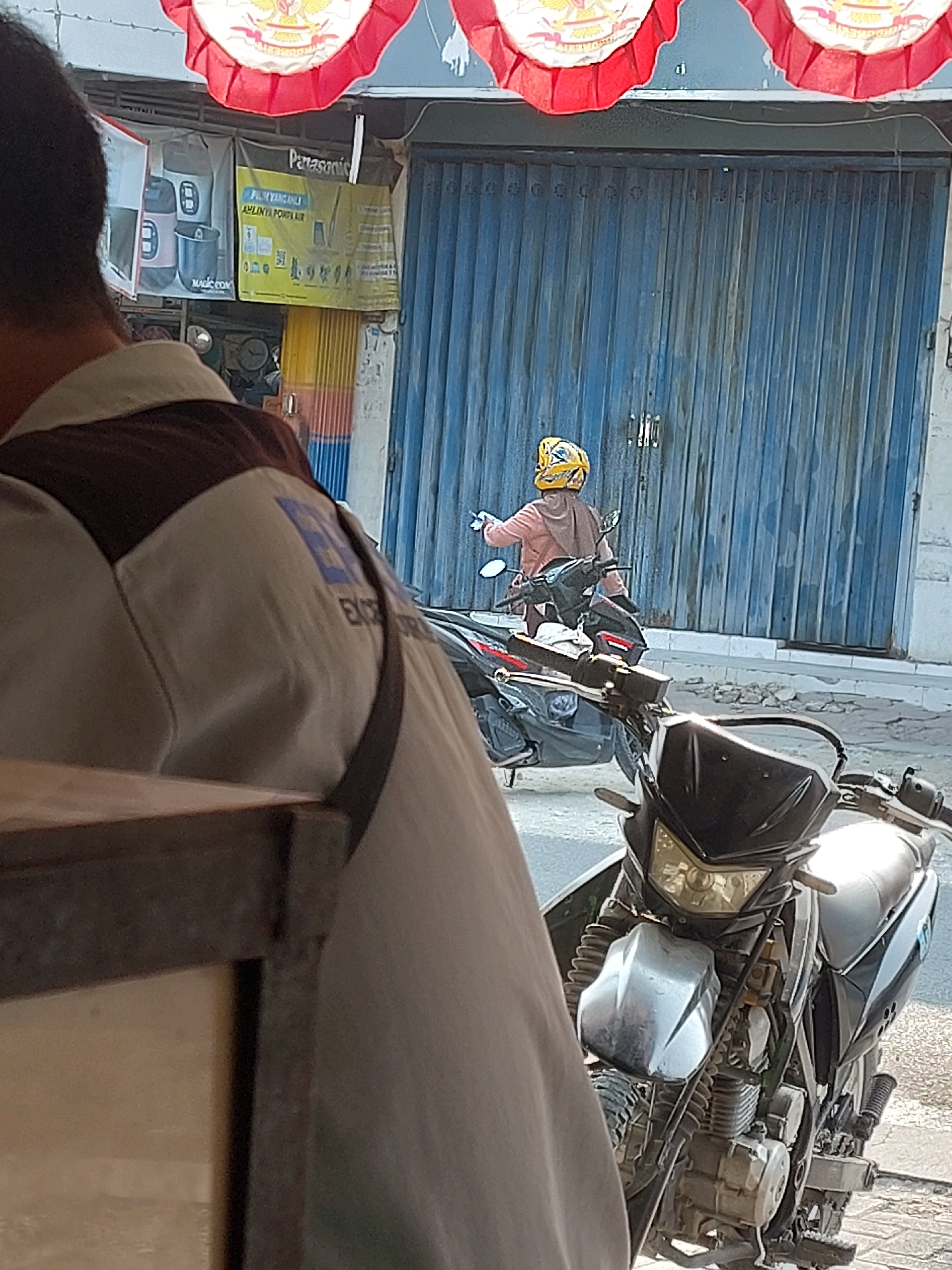 The image depicts a street scene in Sumuragung, Indonesia. In the foreground, partially obscuring the view, is the shoulder and back of a person wearing a light-colored uniform with a dark brown collar and a dark strap across their back. The uniform appears to have a blue emblem on the right shoulder. In front of this person is a motorcycle, predominantly black with silver accents. The motorcycle's front is visible, showing its headlights and handlebars.
In the middle ground, a person wearing a tan headscarf and a light pink jacket is seated on a motorcycle. They are wearing a yellow helmet with a black pattern. This motorcycle is positioned in front of a closed, blue, corrugated metal shutter that covers the entrance to a building. Above the shutter, advertisements are visible, including one that says "Panasonic" in large letters on a yellow background. Red and white decorative banners are also hanging above the advertisements.
The environment appears to be a commercial street with buildings and a paved road. The lighting suggests it is daytime, with sunlight casting shadows. The overall impression is that of a typical street scene in a bustling Indonesian town.