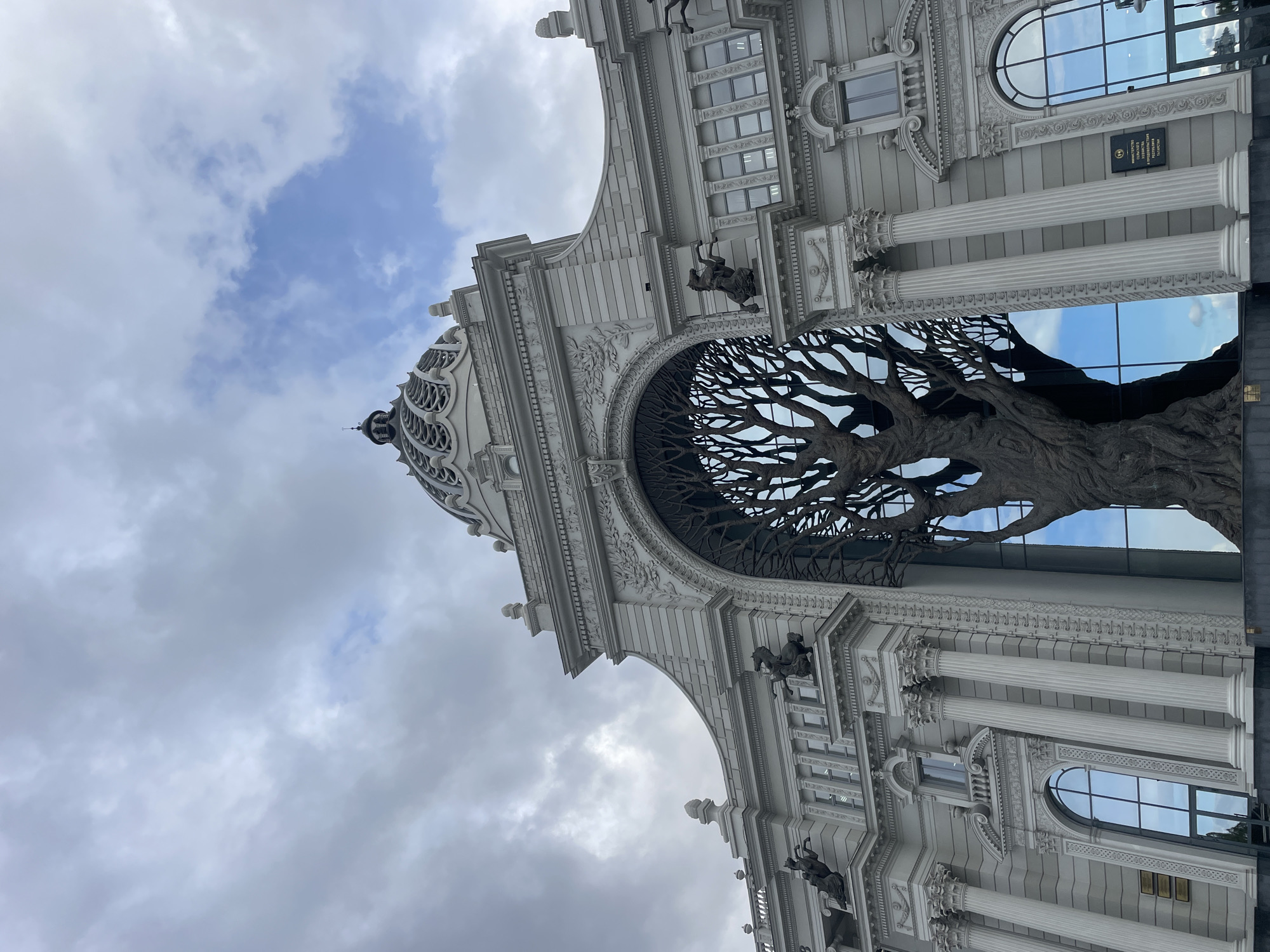 This image captures the grand facade of a stately building, identified as the Ministry of Agriculture and Food of the Republic of Tatarstan, located in Kazan, Russia. The architectural style is classical, featuring light-colored stone or stucco, ornate detailing, and imposing structures.
The most striking feature is a massive, intricate sculpture of a bare, spreading tree, which appears to grow from within a large, central archway of the building. Its dark, gnarled branches fan out, filling the arch and creating a dramatic visual effect.
The building itself showcases fluted columns with detailed capitals, decorative cornices, and arched windows that reflect the sky. Sculpted figures, possibly horsemen, are visible adorning the facade near the archway. A domed section with elaborate openwork is prominent on the upper left side of the structure.
The sky is a mix of overcast grey clouds and patches of bright blue, suggesting daylight conditions. The blue sky is particularly noticeable reflecting in the building's windows and seen through the open branches of the tree sculpture.
A dark plaque on the right side of the building bears white text in Russian: "МИНИСТЕРСТВО СЕЛЬСКОГО ХОЗЯЙСТВА И ПРОДОВОЛЬСТВИЯ РЕСПУБЛИКИ ТАТАРСТАН," which translates to "Ministry of Agriculture and Food of the Republic of Tatarstan." The photograph is taken from a low angle, emphasizing the building's height and grandeur.