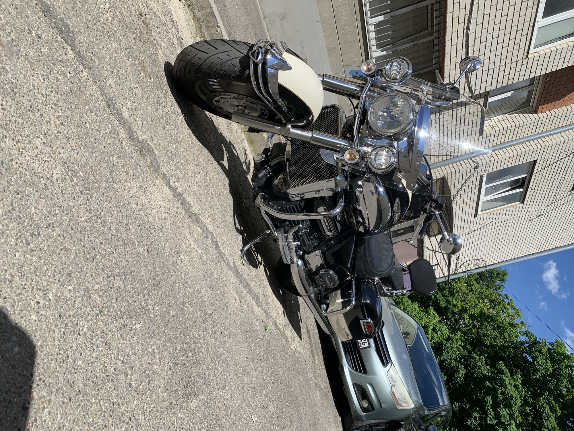 The image captures an outdoor urban scene in Unknown, Unknown, under bright daylight conditions. A large, black and white cruiser motorcycle, identified as a Triumph Rocket III via visible badging on the engine crash bar, is parked prominently in the foreground and midground, angled slightly towards the right. It features extensive chrome detailing, including handlebars, mirrors, front forks, engine guards, exhaust pipes, and multiple headlights. A clear windshield is present, exhibiting a strong sun glare. The motorcycle has a white front fender and black elements for the fuel tank, seat, and saddlebags.
Behind the motorcycle, on the right side of the frame, a silver-green car is parked, partially obscured by the motorcycle. The car's front grille, headlights, and part of its bumper are visible. A Russian license plate, showing "26. RUS" along with other characters, is affixed to the front of the car.
To the left and behind the motorcycle, a multi-story building constructed of light-colored bricks with white-framed windows is visible. A vertical metal pole or conduit runs upwards along the building's facade. In the upper right background, a clear blue sky with sparse white clouds is visible above a dense canopy of green trees.
The ground is a rough, light gray asphalt or concrete surface, showing signs of wear with cracks and textured aggregate. Distinct dark shadows are cast by the motorcycle and potentially other unseen objects, indicating a high sun position. No people are present in the scene.