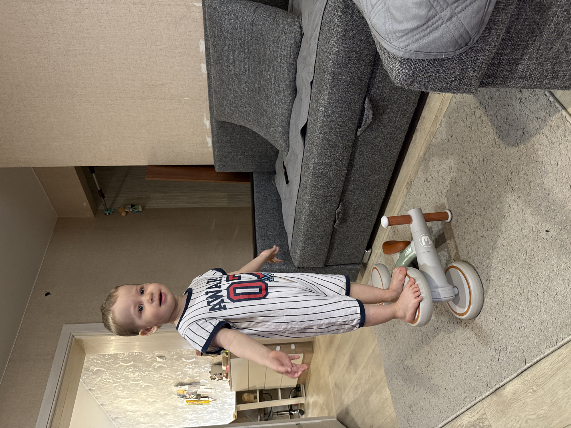 An image captures a young child standing barefoot indoors, looking directly at the camera with a slight smile and curious expression. The child, a toddler with light hair and blue eyes, wears a white and navy blue striped romper. The romper features the text "AWAKE" across the chest in navy blue, with the number "00" (or possibly "OO") in red and blue below it.
The child stands next to a light gray and brown balance bike or ride-on toy, which has "MiniM" visible on its frame. The setting appears to be a domestic living room or similar space. A large, dark gray fabric sofa with quilted armrests and cushions occupies the background to the child's right. The sofa is placed against a light beige wall.
A textured, light gray rug covers the floor where the child and toy are positioned, extending under the sofa. Beyond the rug, a light-colored wooden or laminate floor is visible. In the further background, to the child's left, patterned wallpaper is visible, along with a doorway and faint glimpses of other household items or furniture. The indoor lighting suggests daytime, but no specific time or weather can be discerned. The image's location context is Rusanovo, Russia.