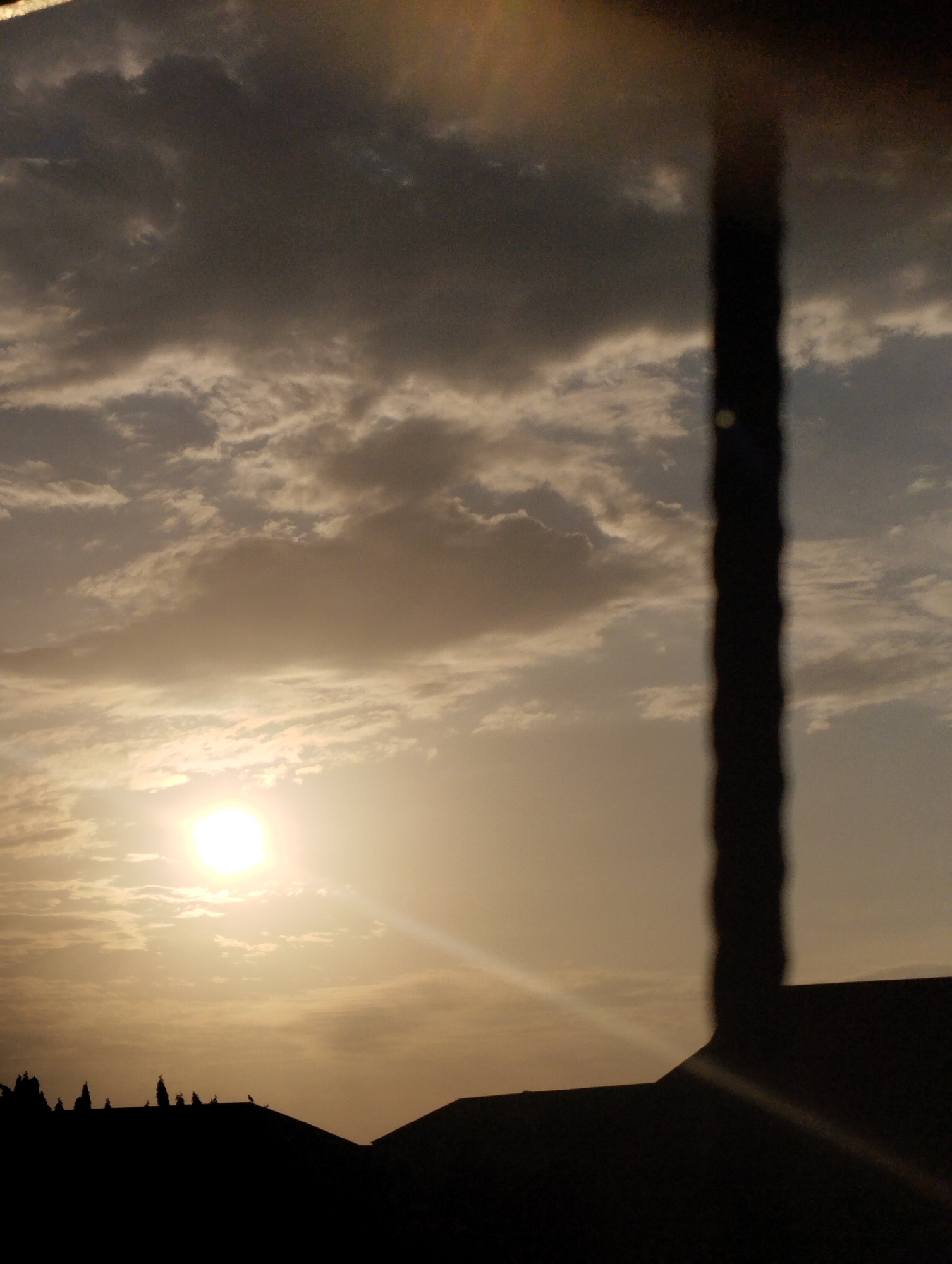The image depicts a sunset scene in Ihiagwa, Nigeria. The sun is low on the horizon, obscured by thin clouds. The sky is a mix of light and dark clouds. In the foreground, there are silhouettes of rooftops and possibly trees. An out-of-focus vertical object appears to be partially blocking the view. There are no people visible in the image. The overall tone is calm.