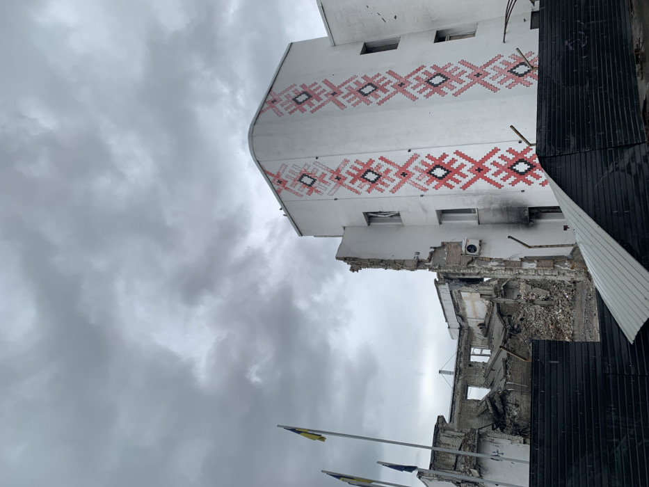 The image depicts a partially destroyed building in Derhachi, Ukraine, located at 50.113 latitude and 36.107 longitude. The building's facade is white with a red and black embroidered design typical of Ukrainian folk art. The top floor of the building appears intact, while the lower floors are severely damaged, with debris scattered on the ground. A black metal fence obstructs the front of the building, and a Ukrainian flag is visible in the background. The sky is overcast, suggesting a recent storm or rain. The scene suggests that the building has been damaged by shelling or other forms of warfare. There are no people visible in the image.