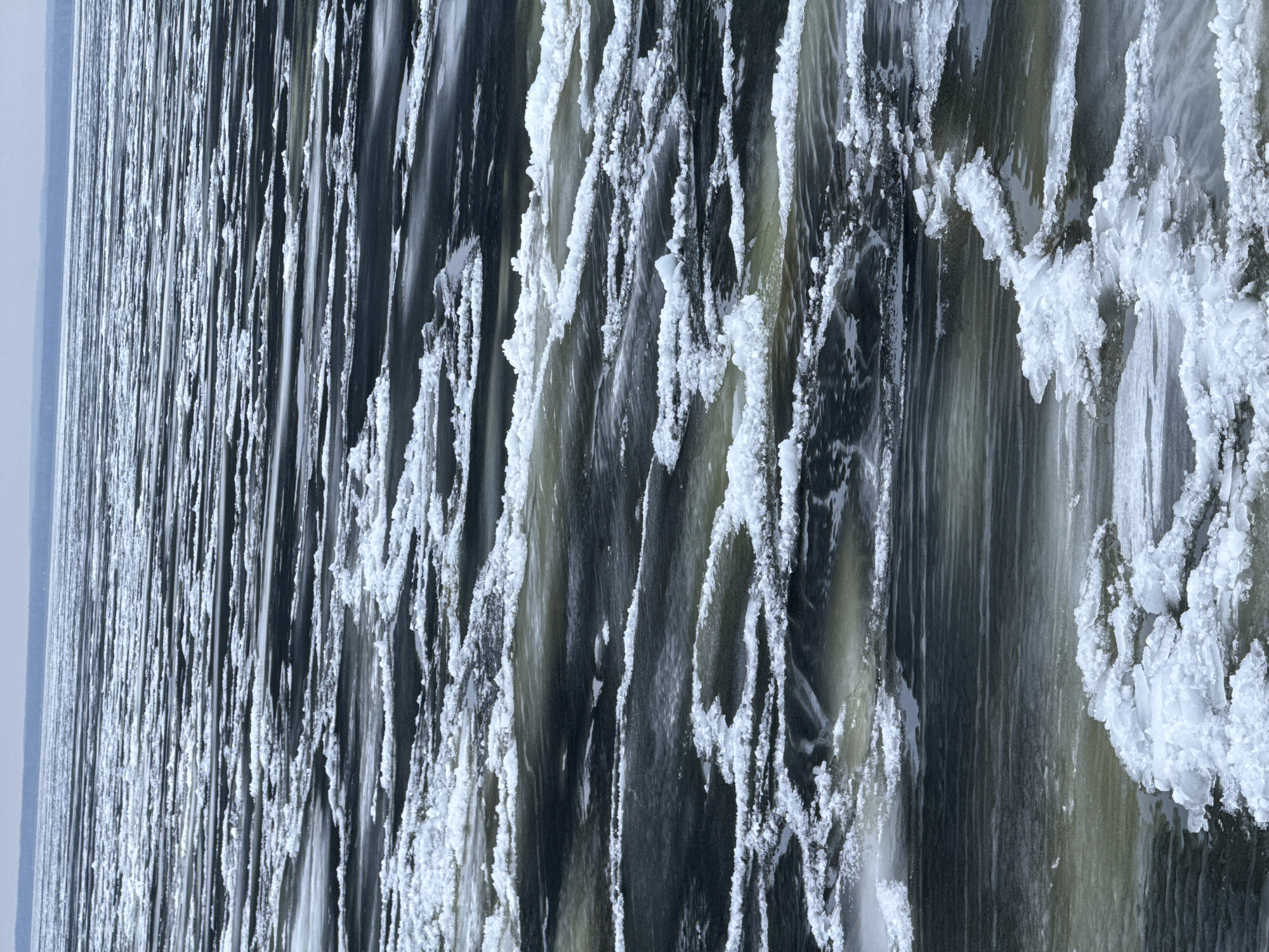 The vertically oriented image depicts a dramatic winter scene of a partially frozen body of water, likely a waterfall or river rapids, characterized by strong contrasts between dark, flowing water and bright, crystalline ice. The setting is outdoors, indicative of cold weather conditions.
The main subjects are the turbulent water and the extensive ice formations. The water, appearing in shades of dark grey, black, and muted greenish-brown, flows vertically downwards with visible streaks and patterns of movement. It appears powerful and fast-moving.
Abundant white ice formations are prominent throughout the frame, clinging to and emerging from the water. These are rugged, irregular, and crystalline, varying from delicate frost-like textures along edges to thicker, chunky icicle-like structures. Some ice formations appear to be actively growing or shaped by the water's flow and spray. The bright white of the ice starkly contrasts with the dark, moving water, emphasizing the freezing temperatures.
In the upper left background, a faint, light grey horizontal band suggests a distant horizon or landmass, adding depth to the scene. The overall lighting is soft and diffused, typical of an overcast winter day, with no harsh shadows. No text is visible in the image. The scene, potentially located in Chaikovsky, Russia, conveys a sense of raw natural power and the extreme beauty of a frozen landscape.