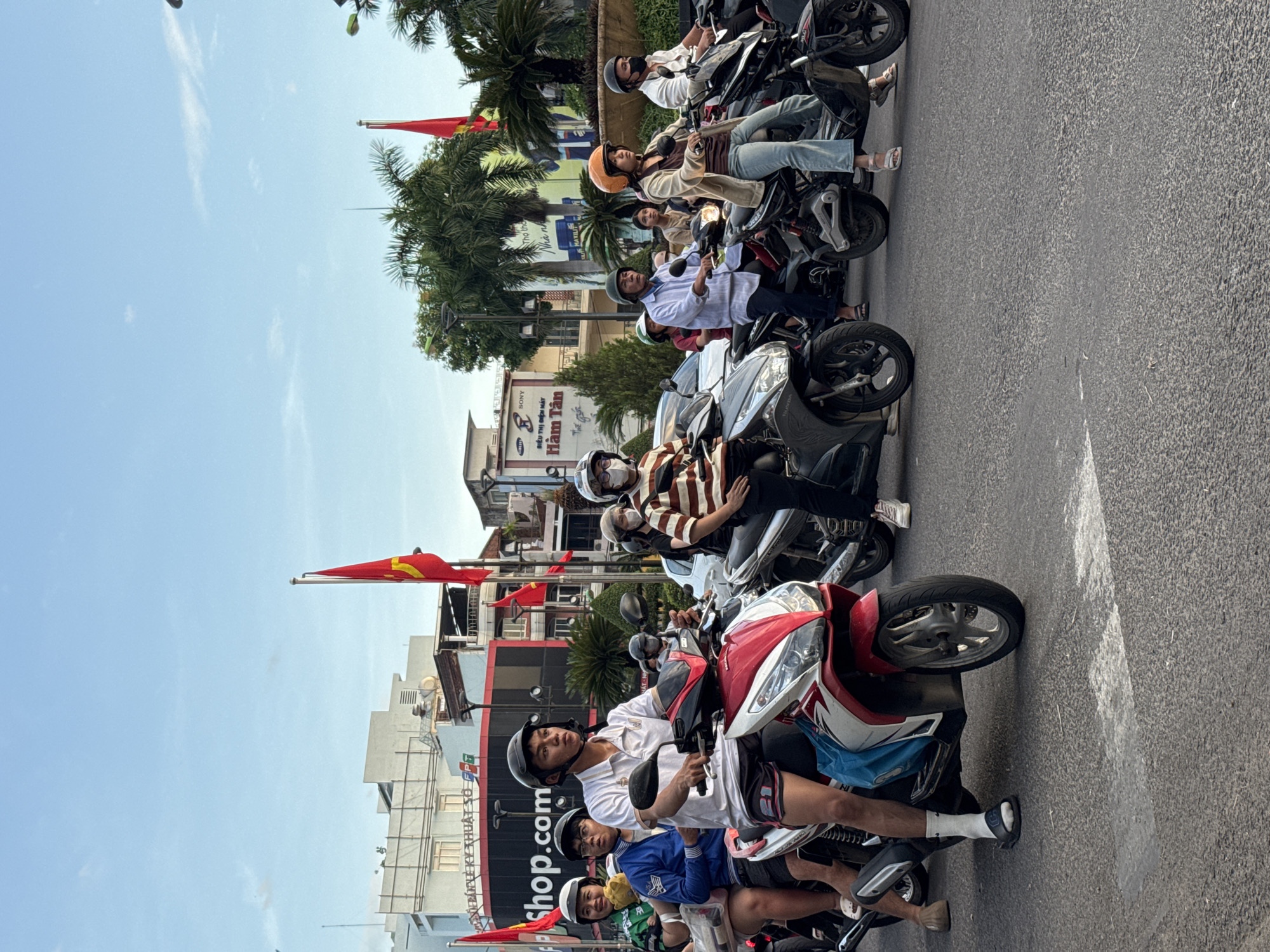 This image depicts a street scene in Nha Trang, Vietnam, likely during the daytime with overcast skies. Several individuals are riding motorcycles in what appears to be traffic.
In the foreground, a young man wearing a white polo shirt, black shorts with a red stripe, and a black helmet is riding a red and white motorcycle. He is wearing white socks and black sandals. He is looking forward, seemingly engaged in riding. Behind him on the same motorcycle, a woman with glasses and a green shirt is seated, holding a light-colored object. Beside them, another person in a blue jacket and a helmet is also on a motorcycle.
Further in the background, other motorcycles and riders are visible. One motorcycle has a woman with an orange helmet wearing a light brown jacket and blue jeans, looking to the left. Another motorcycle has a rider in a striped shirt and a white helmet. Several people are wearing helmets and masks, suggesting a common practice for safety or health reasons.
The environment includes buildings in the background, some with signage that is partially visible, suggesting a commercial area. Palm trees are also present, indicative of a tropical or subtropical location. Red flags with yellow stars, likely Vietnamese flags, are displayed on poles along the street, along with smaller flags. The road surface is asphalt with some visible lane markings. The overall impression is a busy urban street with common modes of transportation.