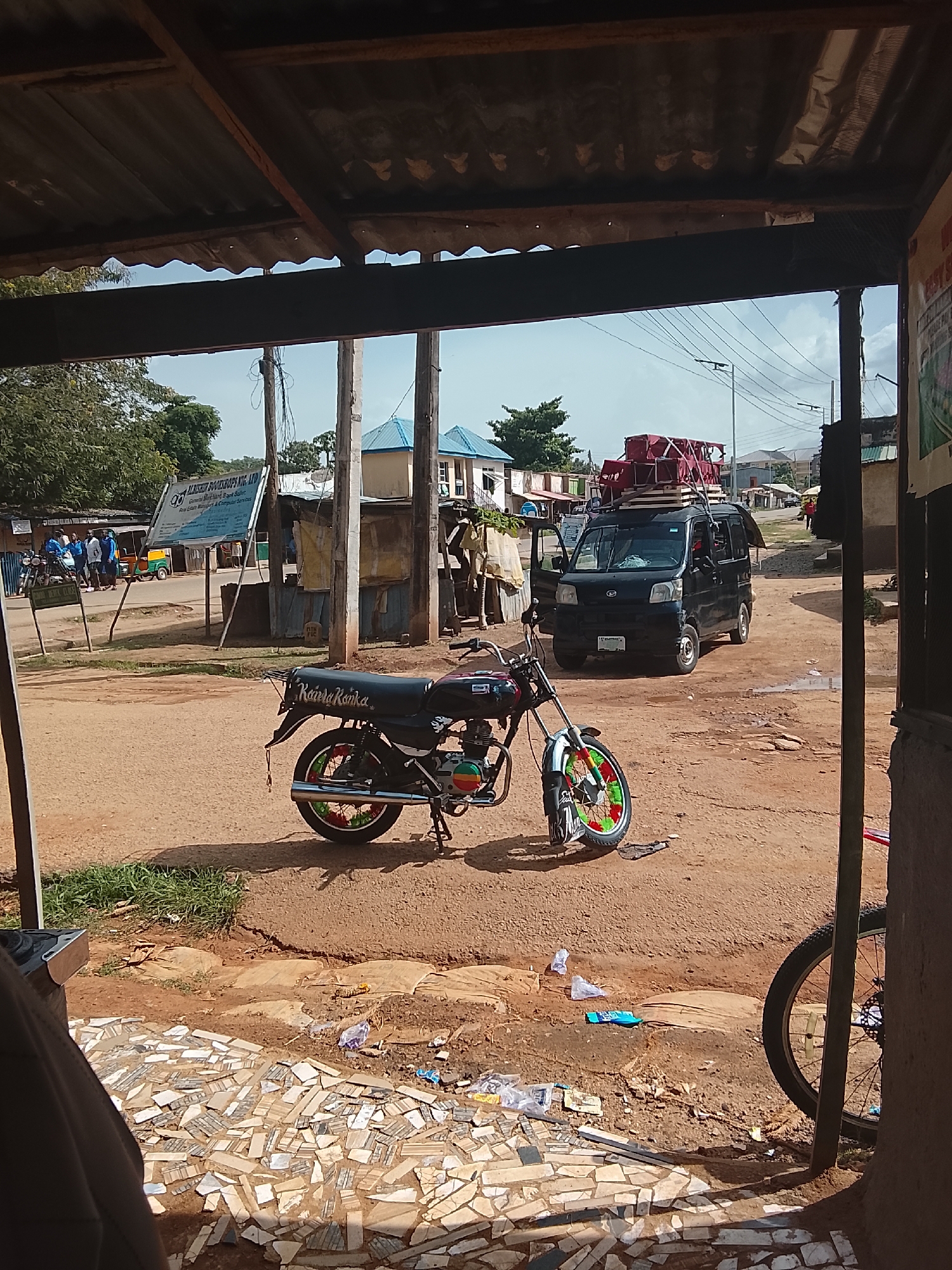 The media file depicts an outdoor scene, likely in a rural or semi-urban area, bathed in daylight. The foreground is dominated by a motorcycle with a black body, decorated with vibrant red, green, and yellow spokes on its wheels. Its license plate is not clearly legible. In the background, a black van is parked, its rear cargo area piled high with red crates and other items, suggesting transport or goods delivery. The van's doors are open.
Several people are visible in the middle ground. On the left, a group of individuals, some wearing blue attire, are gathered near a signpost. Further to the right, more people are seen near buildings. A three-wheeled vehicle, commonly known as an auto-rickshaw, is also present on the left side of the scene. The setting appears to be a marketplace or a roadside stop, with various structures made of corrugated metal and wood. A large sign with text is mounted on a post, but the text is difficult to read clearly from this perspective. A bicycle's wheel is visible on the far right. The ground is unpaved and appears to be a mix of dirt and scattered debris. The overall impression is one of an active, everyday scene in a location where transportation and commerce are taking place. The sky is visible in the upper portion, indicating a daytime setting with what appears to be fair weather.