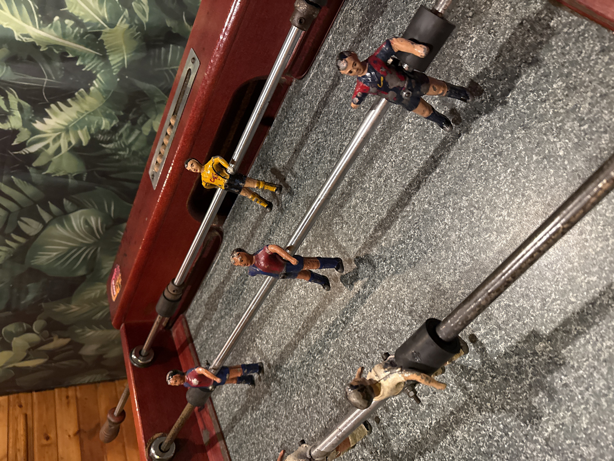 The image shows a close-up of a foosball table. The table is red with chrome rods that hold small, painted soccer player figures. The figures wear different colored jerseys. The playing surface is gray with a speckled pattern. The background includes a green and white patterned wall, possibly wallpaper, and a glimpse of wooden paneling. The image does not provide enough information to determine the location in Barcelona.