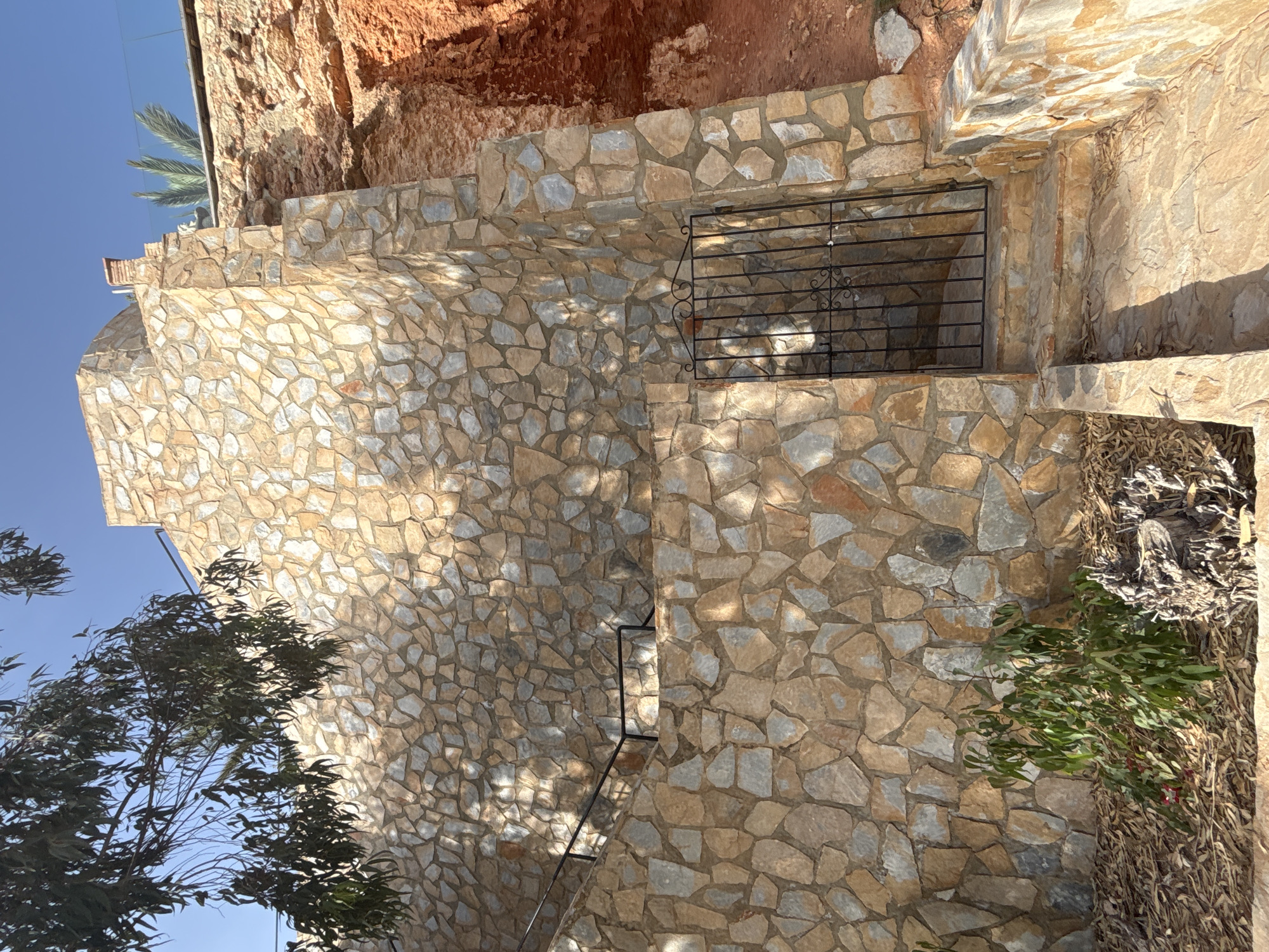 The image shows a stone wall with a black metal gate and stone steps leading up to it. The wall is constructed from irregularly shaped stones in shades of tan, grey, and light orange. To the right of the wall is a cliff face of reddish-brown rock. A small green bush is planted at the base of the wall, and dried leaves are scattered on the ground. Above the wall, a bright blue sky is visible, with the branches of a tree with green leaves hanging into the frame from the left. In the background, on top of the stone wall, a palm frond and a section of a glass railing can be seen, suggesting a terrace or elevated area. The scene appears to be an exterior view of a building or property in Urbanización Cabo Roig, Spain.