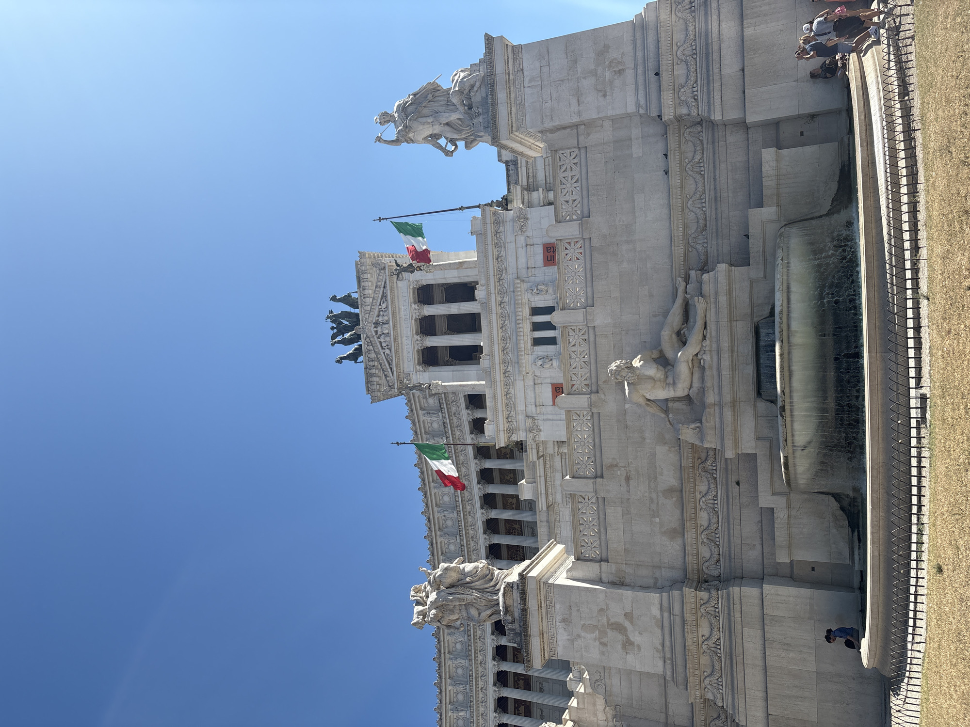 The image shows the Altare della Patria, also known as the Victor Emmanuel II National Monument, located in Rome, Italy. The monument is made of white marble and features several statues and architectural elements. In the foreground, there is a fountain with water flowing. In the middle ground, there are people gathered around the monument and the fountain. In the background, the clear blue sky is visible. The monument is adorned with Italian flags.