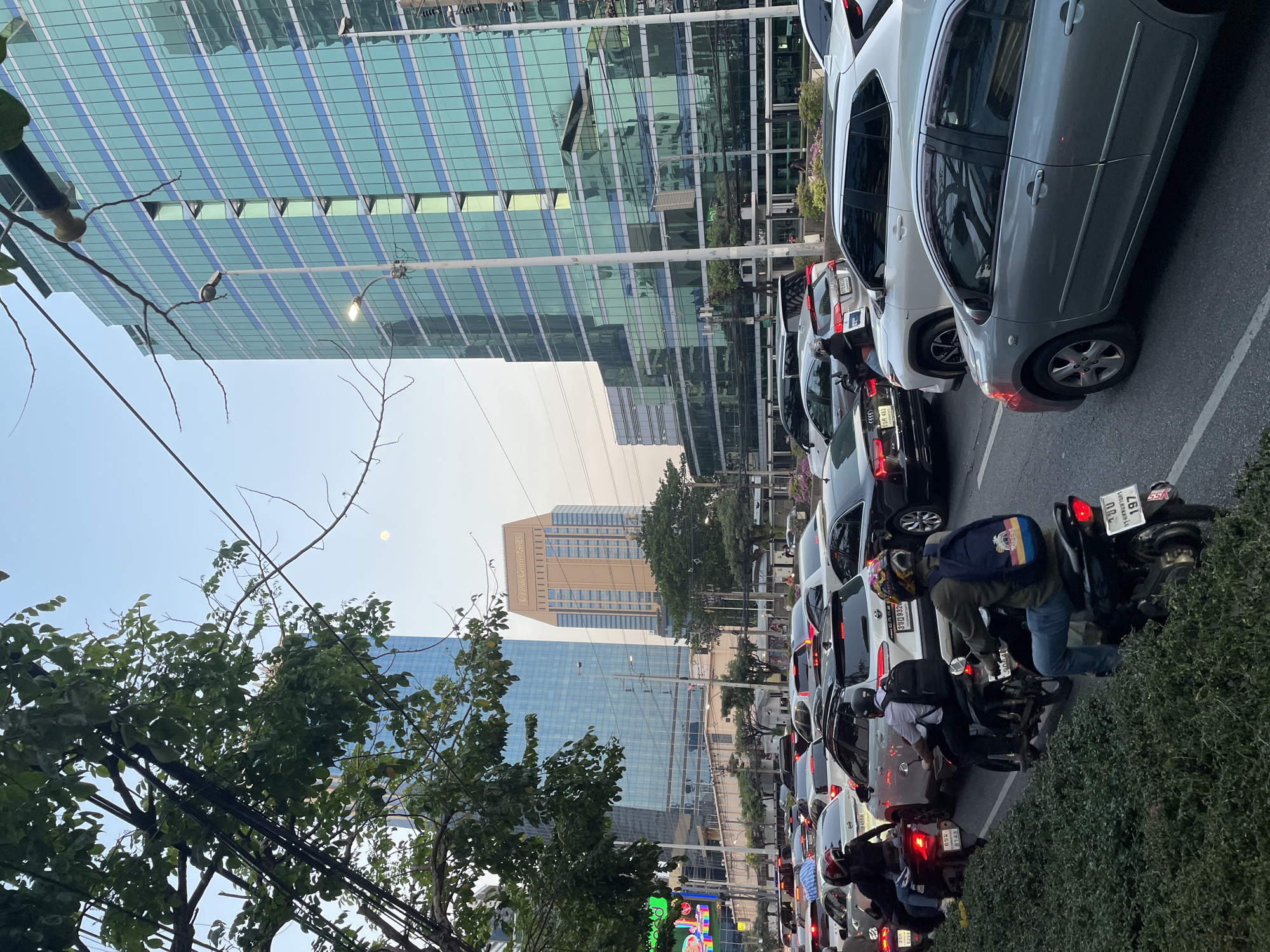 This media file captures a bustling urban street scene in Bangkok, Thailand, likely during the late afternoon or early evening. The sky is a clear light blue, with a pale moon subtly visible. The foreground and midground are dominated by heavy traffic, featuring numerous cars (sedans and SUVs) and motorcycles moving slowly or stopped on a multi-lane road.
Modern high-rise buildings with glass facades form the backdrop, reflecting the sky and surrounding structures. A prominent beige building further down the street clearly displays the text "Grand Centre Point." Several people on motorcycles are seen navigating through the congestion; one individual wears a blue backpack and helmet, while others also wear helmets. Green foliage from trees and a network of utility lines are visible on the left and framing the scene.
Notable details confirming the location as Bangkok include the specific "Grand Centre Point" landmark and the Thai script present on vehicle license plates. Visible text from license plates includes "3ขฎ 9326," "ขย 4559," and "กรุงเทพมหานคร" (Bangkok), along with partial numbers like "5298," "8143," "197," and letters "SSA." A bright sign at the bottom also shows the text "Pomt." The scene conveys the typical vibrant and congested atmosphere of a major city.