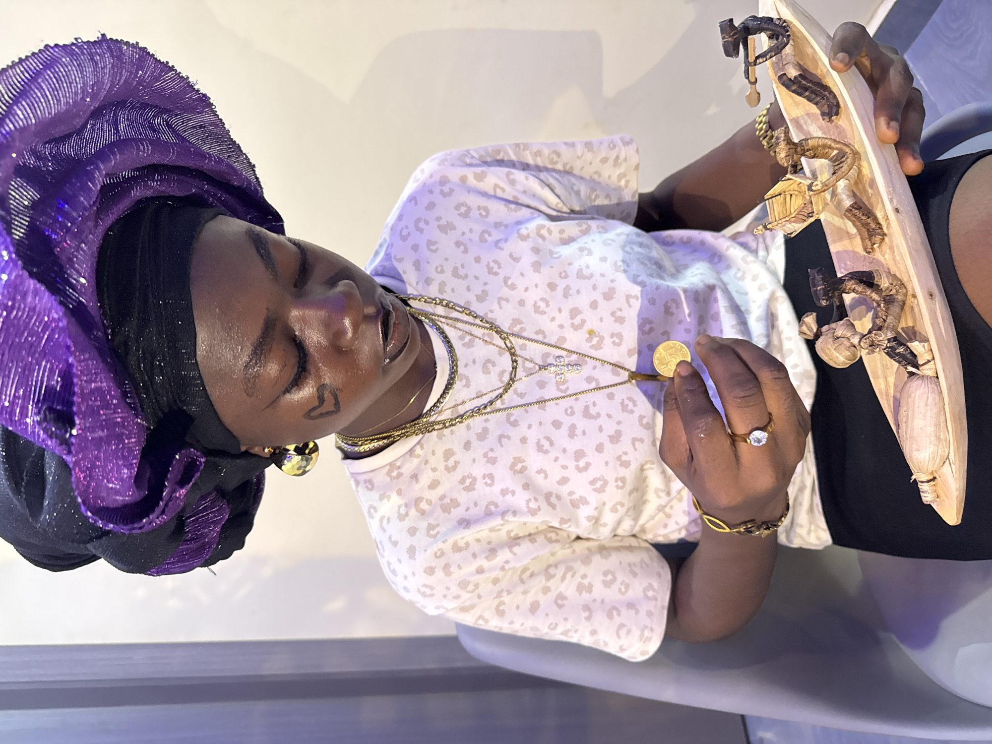 The image depicts a person wearing a purple and black headwrap, several gold necklaces, and a light-colored animal print shirt. The person has a small heart drawn on their face. They are holding a gold coin in one hand and a long, light-colored object with small, dark figurines attached to it in the other hand. The background appears to be a room with a light-colored wall. There is no information in the image to support this being taken in Ajara, Nigeria.