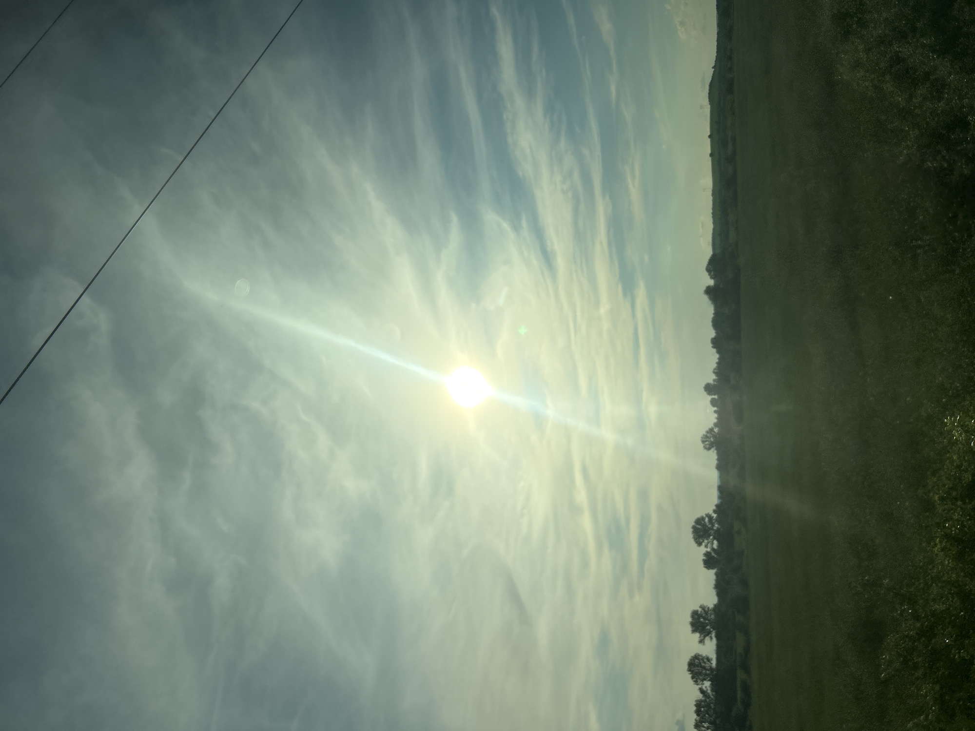 The image shows a landscape with a bright sun shining through wispy clouds. A power line runs diagonally across the sky. Below the sky is a green field with a line of trees and bushes along the horizon. In the far distance, there are rolling hills with some structures that appear to be utility poles. The overall scene suggests an outdoor, rural environment bathed in natural light. The location is identified as Kochovo, Bulgaria.