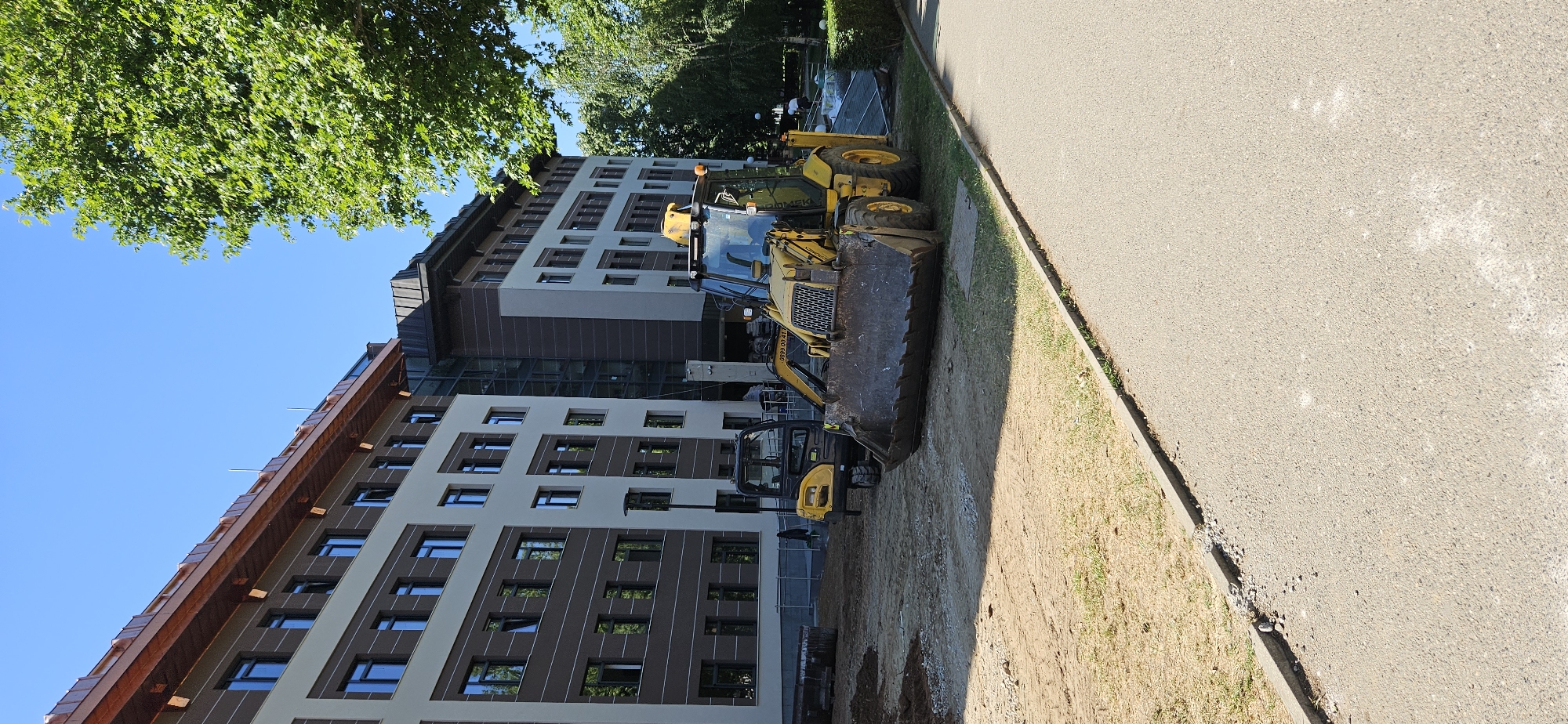 The image shows a construction site in front of a modern building in Blagoevgrad, Bulgaria. The building has multiple floors with brown and beige facade panels and many windows, some reflecting the sky. In the foreground, a yellow backhoe loader with a large front bucket is parked on an area of dirt and disturbed ground next to a sidewalk. A smaller yellow construction vehicle sits behind the backhoe. The sidewalk is gray and runs along the right edge of the image. Lush green trees partially obscure the building and cast shadows on the site. The sky is clear and blue. The overall impression is that construction or landscaping work is in progress.
