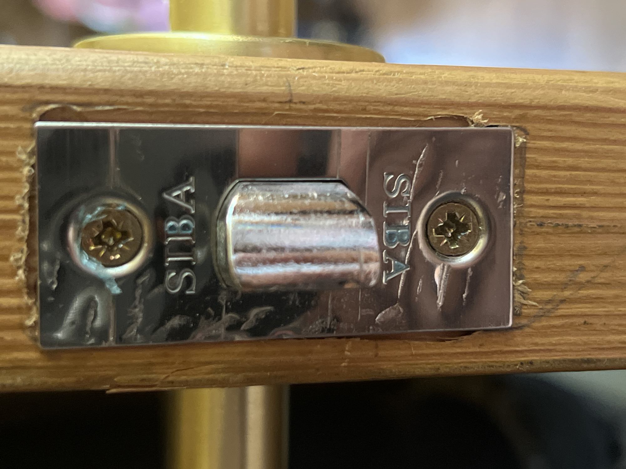 The image shows a close-up, head-on view of a door latch mechanism embedded in a wooden door frame. The latch plate, made of polished metal, is rectangular and secured by two Phillips head screws. The brand name "SIBA" is embossed twice on the metal plate, once upright and once inverted, suggesting it is a reversible mechanism. The latch bolt itself is visible in the center, a shiny cylindrical piece that extends from the plate. The wooden door frame surrounding the latch plate shows signs of wear and tear, with small splinters and indentations. The background is out of focus, but hints at indoor lighting and possibly other objects. The location is identified as Norviliškės, Lithuania.