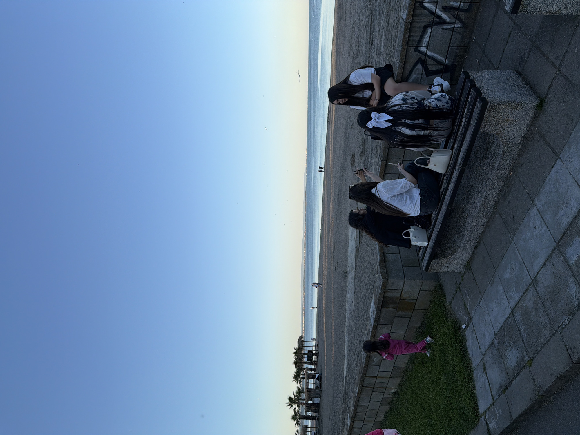 The image, correctly oriented, captures a clear late afternoon or early evening scene at a beachside promenade. A vast, clear blue sky dominates the upper portion, meeting a sandy beach and the calm sea in the middle ground. The foreground on the right features a paved walkway, benches, and a strip of grass.
Four young women are seated on two dark-colored benches. On the farther bench, one woman with long dark hair, wearing a white top and dark bottoms, leans forward. Next to her, another woman with long dark hair tied with a prominent white bow wears a patterned top. On the closer bench, one woman in a white top and dark bottoms holds a mobile phone, and a white handbag rests beside another person (partially visible) in dark attire. They appear to be relaxing and enjoying the view.
On the green grassy strip to the right, a small child dressed in a pink tracksuit stands and looks towards the beach. The paved area consists of dark grey square tiles, and a low stone wall with visible graffiti, including a letter "W," runs behind the benches. In the distance along the beach, several small figures are walking, and palm trees are visible along the coastline. The sea appears calm under the soft, clear light. No other discernible text is visible.