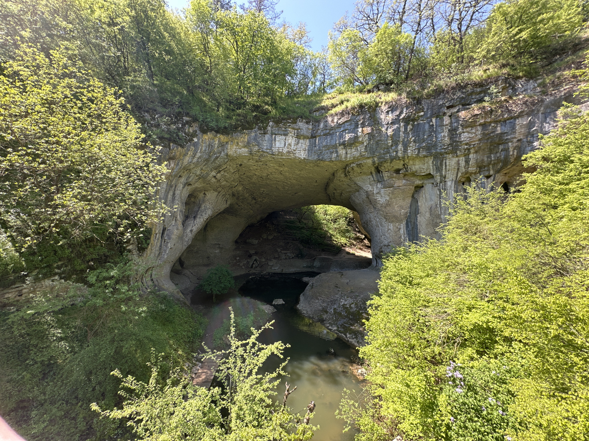 The image shows a natural stone bridge in Lilyache, Bulgaria. The bridge is a large, arched rock formation with a river flowing beneath it. Green trees and vegetation cover the top of the bridge and surround the area. The river is calm and reflects the sunlight. The sky is clear and blue.