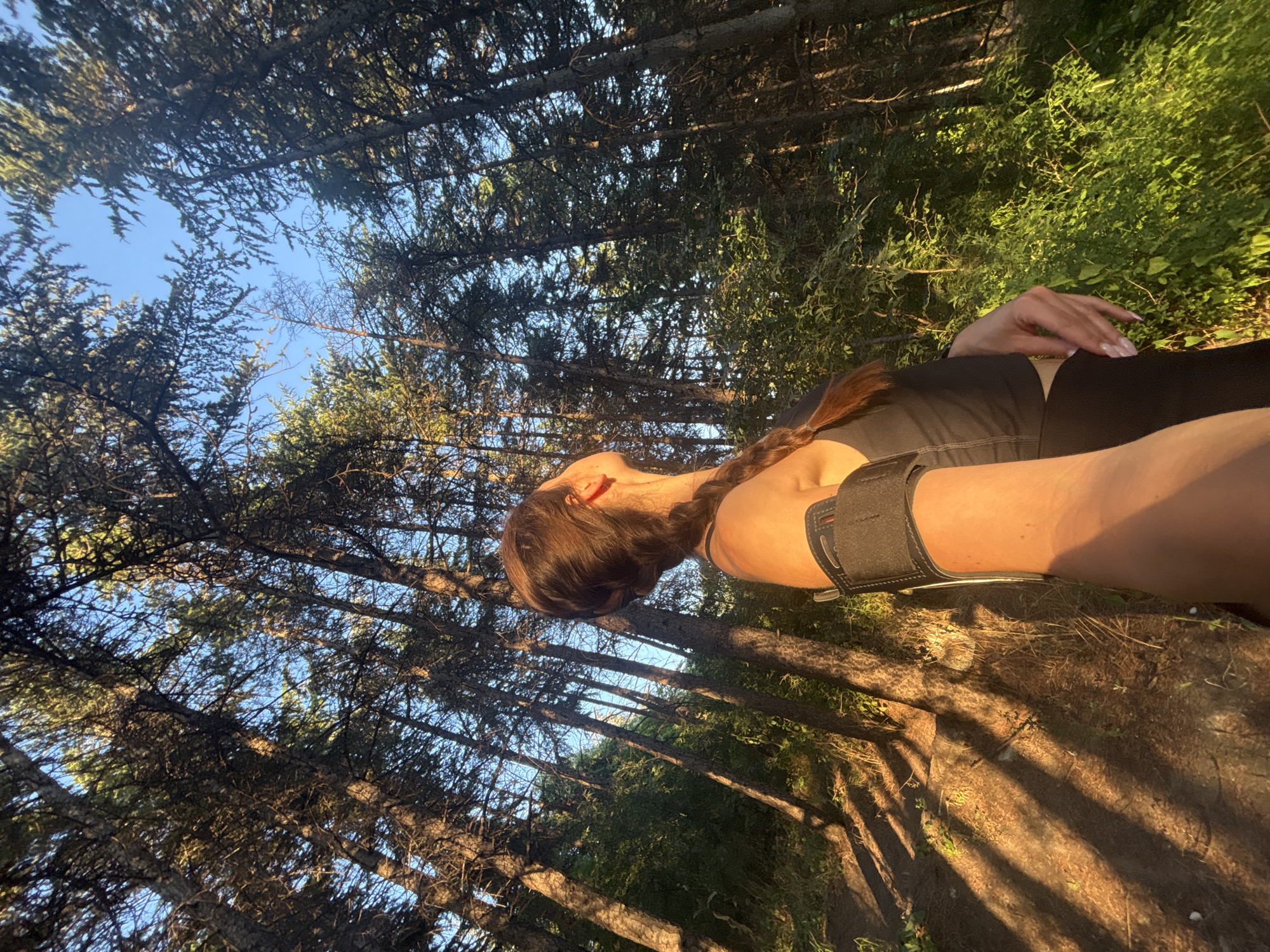 The image shows a person lying down in a forest with their head tilted back, looking up at the sky. The person has long, dark hair braided into a single plait. They are wearing a black sports bra and black leggings, and have an armband on their left arm. The forest appears to be dense with pine trees, and the ground is covered in dry pine needles and some fallen branches. Sunlight filters through the trees, casting dappled shadows on the ground and illuminating parts of the scene with a warm, golden light. The sky is a clear blue, visible through the gaps in the tree canopy. The time of day appears to be late afternoon or early evening, given the quality of the light. The overall mood of the image is peaceful and serene, suggesting a moment of rest or relaxation in nature. The location is identified as Varna, Bulgaria. There is no visible text in the image.