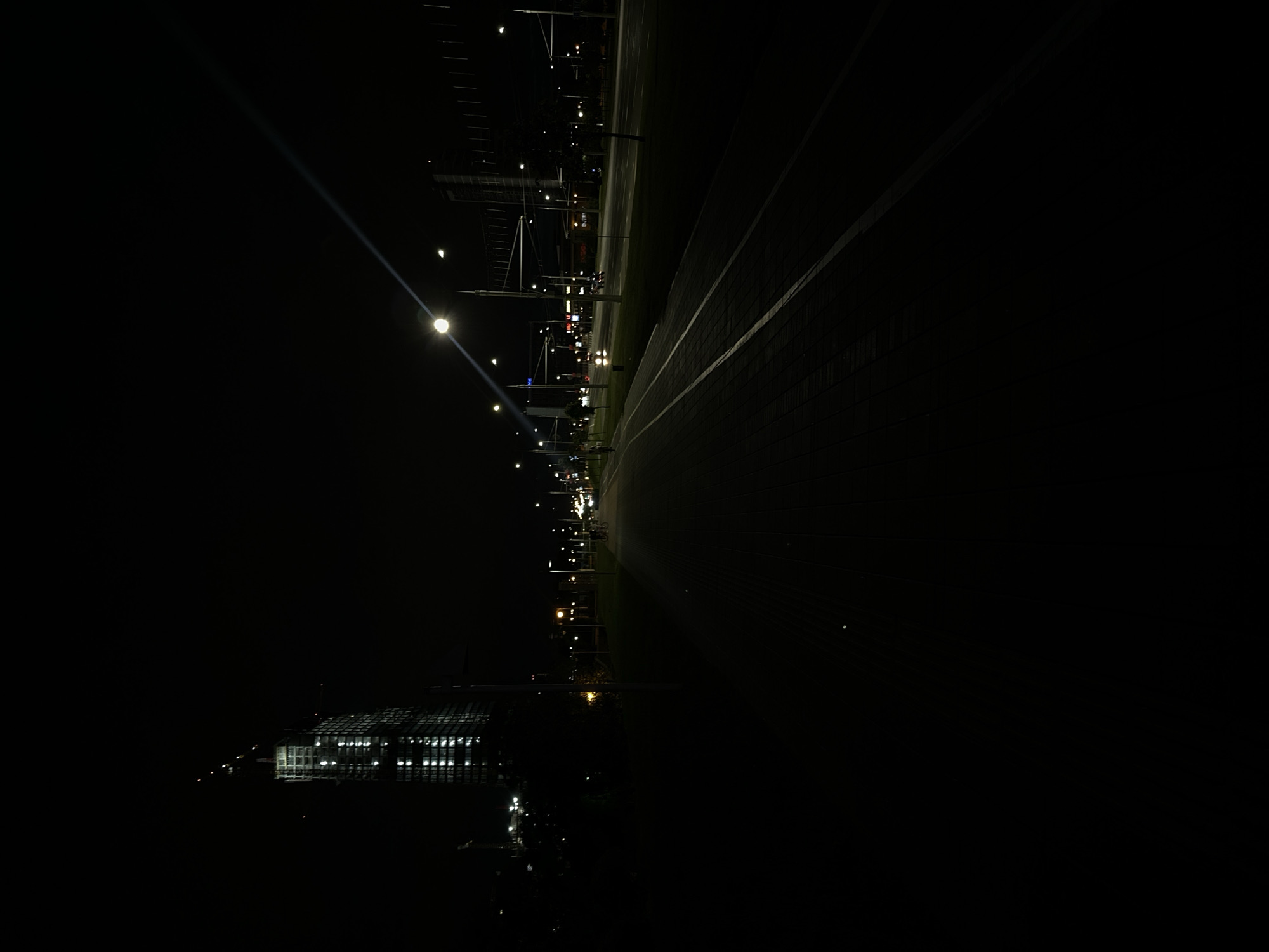 The image depicts a street scene at night in an urban environment. A tall, under-construction building with illuminated windows is visible on the left side of the frame, along with construction cranes. A lamppost casts a bright light on the right side of the image, creating a strong glare and a light beam extending diagonally across the dark sky. The foreground shows a paved walkway with distinct lines, possibly a pedestrian path or bike lane, adjacent to a grassy area. Across the street, there are more streetlights, buildings with illuminated signs, and the headlights of moving vehicles. Several people are visible on the walkway in the middle distance, some appearing to be on bicycles. The overall atmosphere is dark and illuminated by artificial lights, suggesting a typical urban night.