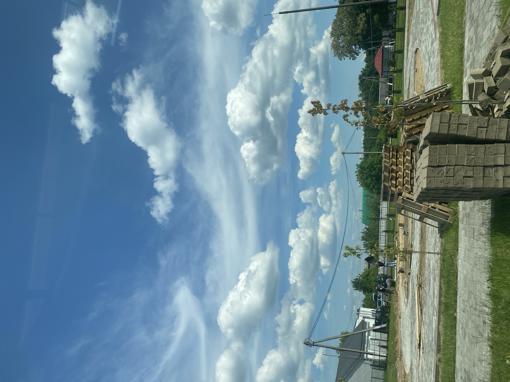 The image depicts a construction site under a partly cloudy blue sky. In the foreground, stacks of paving stones and wooden pallets are visible. The ground is covered with paved walkways and areas of sand, suggesting the early stages of landscaping or public space development. In the middle ground, several individuals appear to be engaged in construction work, possibly operating a cement mixer. A small tree stands near the center of the frame, with other trees and buildings forming the background. The overall scene suggests an outdoor construction project taking place in a suburban or semi-rural setting within the city of Krupki, Belarus.