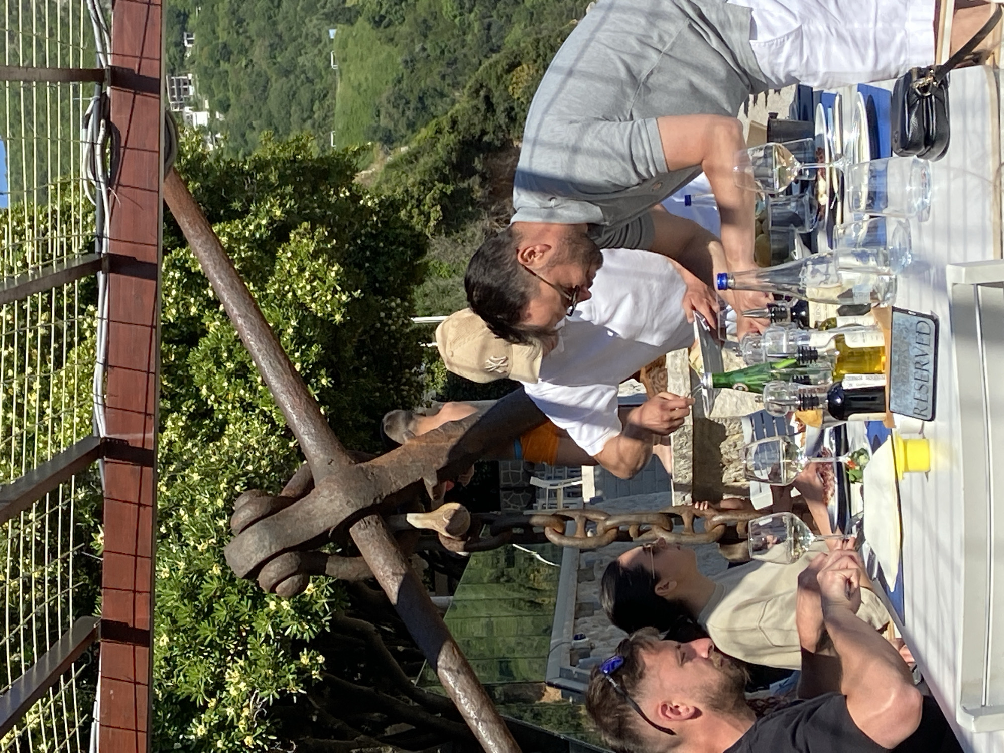 The image shows a group of people having a wine tasting at an outdoor venue, possibly a restaurant or vineyard, in Prijevor, Montenegro.
The scene includes a table with wine bottles, glasses, a "Reserved" sign, plates with food (possibly appetizers or tasting portions), and a black purse on the right side. People are gathered around the table, tasting wine and socializing.
The location appears to be a scenic spot with a view of the surrounding hills and vegetation. An iron anchor is positioned prominently in the foreground. There's a fence structure to the left, beyond which the hills of Prijevor are visible.