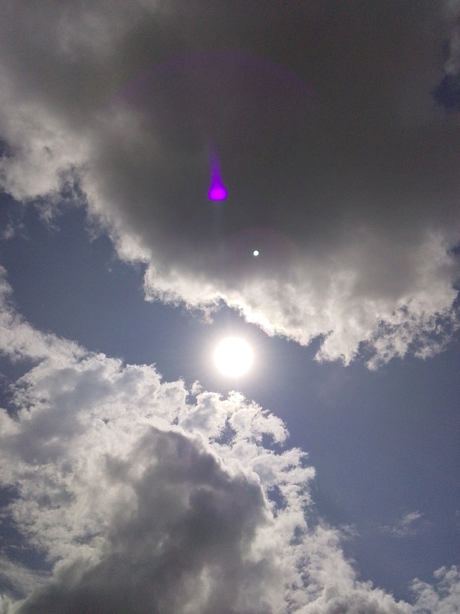 The image shows a view of the sky from Serti, Nigeria, with several clouds obscuring parts of the sun. A bright, circular sun is visible in the lower right quadrant of the image. There is a purple light anomaly present in the upper portion of the image, possibly a lens flare or reflection. The sky is blue, with scattered white and grey clouds. The image is taken from a low angle, looking upwards. There are no people or man-made objects clearly visible in the frame, indicating a natural outdoor scene.