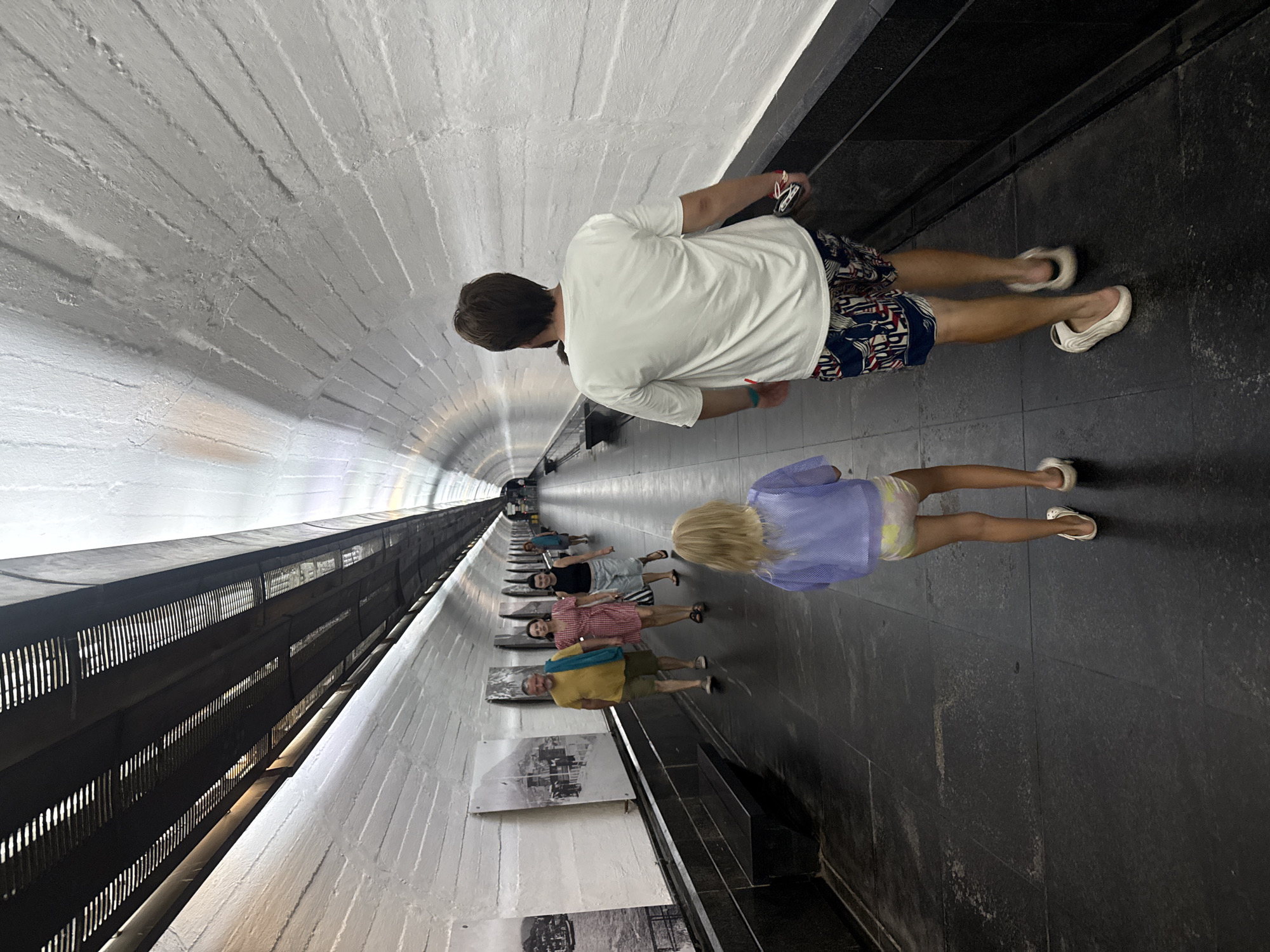 A group of people are walking along a dark, tiled pathway inside a long, arched tunnel in Yalta, Ukraine. The tunnel walls are textured and painted white, with some artwork displayed on the left side. A man, a woman, and a child are in the foreground, with other people further down the tunnel. The man in the foreground is wearing a white t-shirt and patterned shorts, walking with a young girl in a blue jacket and shorts. The girl is walking slightly ahead of him. The man is carrying a dark object in his right hand. The pathway is illuminated by overhead lights that cast long shadows.
