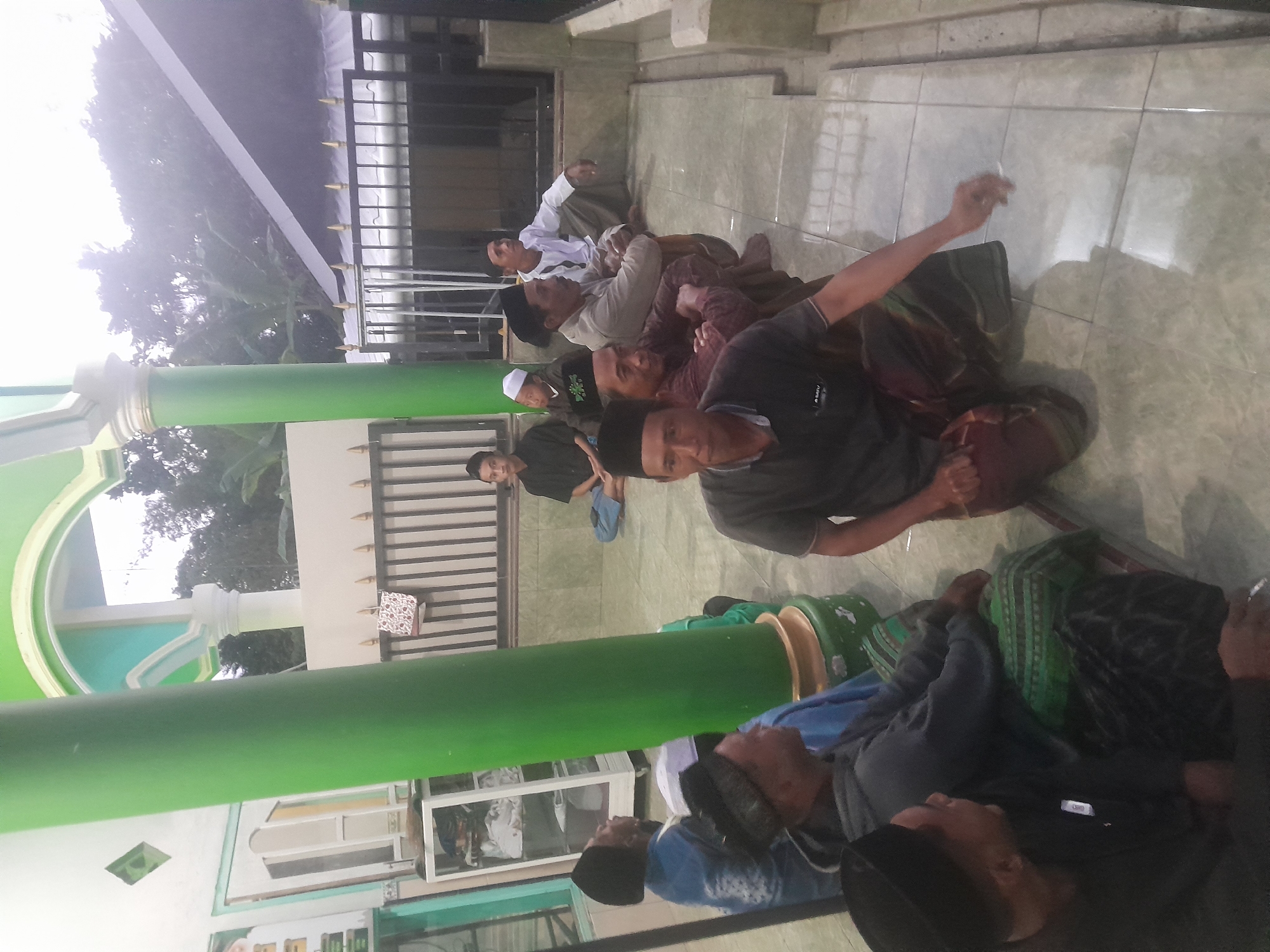 In the city of Parijatah Kulon, Indonesia, a group of nine adult males are gathered outdoors near a mosque. The scene is captured from a slightly low-angle perspective, focusing on the individuals seated on the tiled ground. The mosque features a prominent green pillar and a green-trimmed architectural element overhead. In the background, a black-tiled roof slopes downwards, partially obscuring the sky, which appears overcast. A black metal fence separates the mosque area from the background.
The men are dressed in traditional Islamic attire, primarily consisting of sarongs and caps. Three men are in the foreground, with the closest one in a black t-shirt and a patterned sarong, looking towards the left. To his right, another man in a maroon and black patterned shirt and a darker sarong is also seated. Further back and to the left, a man in a grey hooded jacket and a green and black sarong is partially visible.
Behind them, several other men are seated in a row. One man in a black t-shirt and a white cap is visible on the far left. Next to him, a man in a light brown shirt and a patterned sarong is seated, looking towards the right. Another man in a white shirt and a dark sarong is seated to his right, appearing to gesture with his hand. Further back, two men in dark clothing are seated.
The environment appears to be an outdoor gathering space associated with a religious institution. The overall atmosphere is calm and communal, with the men appearing to be engaged in conversation or religious observance.