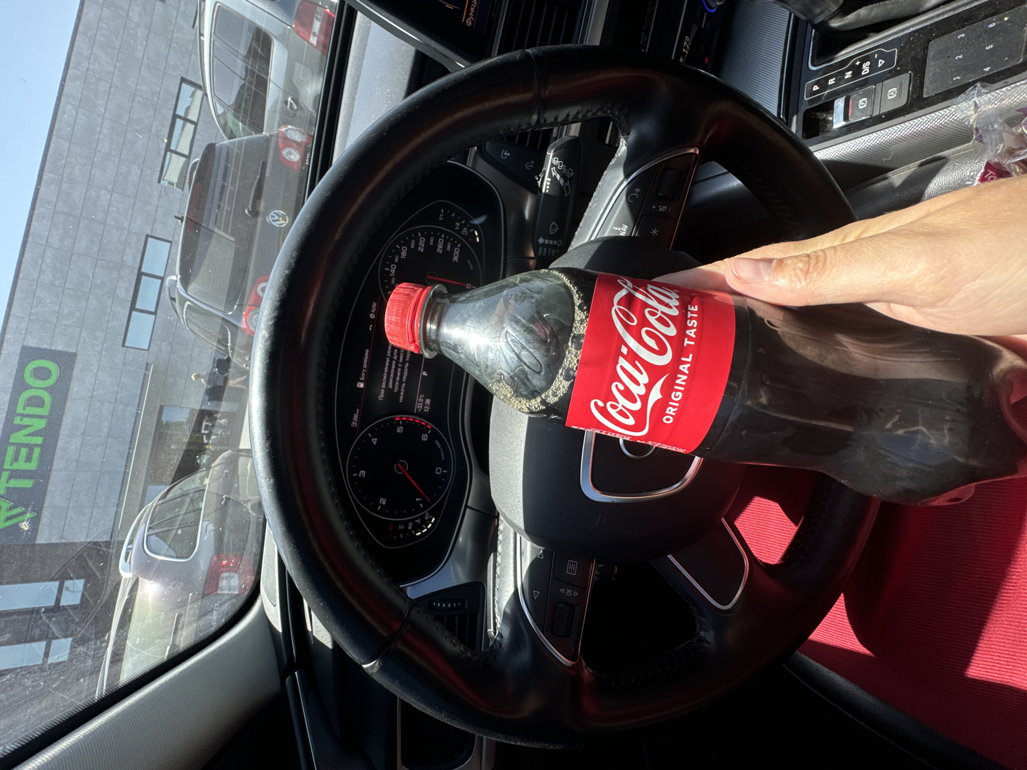 The image shows the interior of a car, viewed from the driver's perspective. A hand is holding a bottle of Coca-Cola in front of the steering wheel. Through the car's windshield, a parking lot is visible, with several cars parked and a building in the background. The building has a green sign with the word "TENDO" on it. The license plate of one of the visible cars is blurred. The car's dashboard and steering wheel are clearly visible. The image appears to have been taken in the city of Sarny, Ukraine. The presence of the "TENDO" sign on the building suggests it may be a store or shopping center in that city.