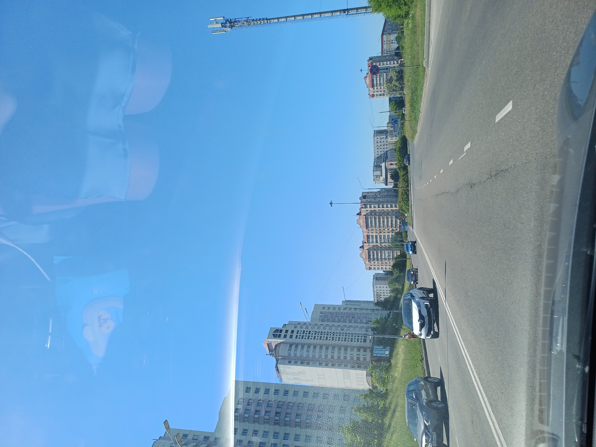 The image depicts a daytime scene of a city street with apartment buildings lining the right side of the road. The sky is clear and blue, reflecting some shapes on the windshield of the vehicle from which the photo was taken. Several cars are driving on the multi-lane road. A tall cell tower is visible in the distance on the right side of the image. The perspective suggests the photo was taken from inside a moving vehicle.
