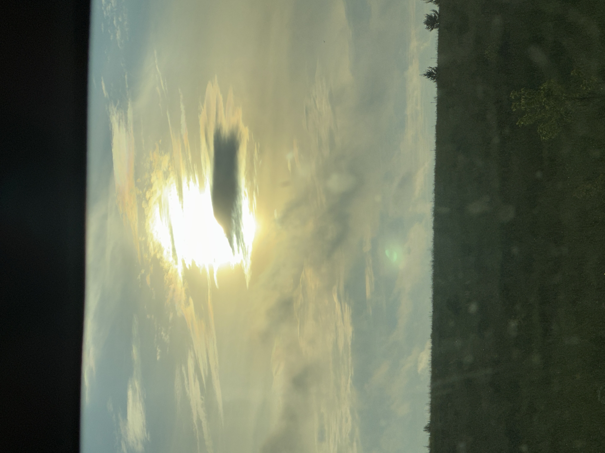 A wide shot captures a cloudy sky at sunset over a grassy hill in Buranovo, Russia. The sun is partially obscured by clouds, casting a warm glow across the sky with hues of yellow and orange. The foreground is dominated by a dark, grassy field, with a few scattered trees and bushes. The overall scene conveys a sense of natural beauty and tranquility.