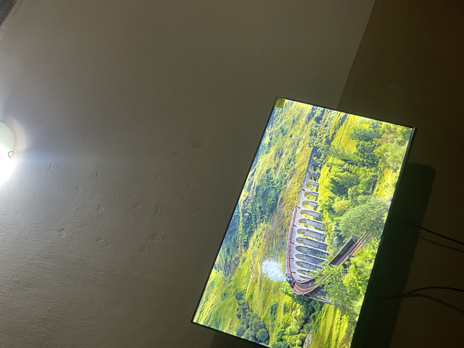 A television screen dominates the scene, displaying a vibrant landscape of a train traversing a viaduct amidst lush greenery. The scene evokes a sense of adventure and travel, perhaps familiar from popular culture. A dim, possibly indoor setting surrounds the screen. A wall with imperfections and a light fixture are visible on the left, while a darker wall appears on the right, both indicating the room's ambiance. The angle of the shot suggests an informal, personal setting.
