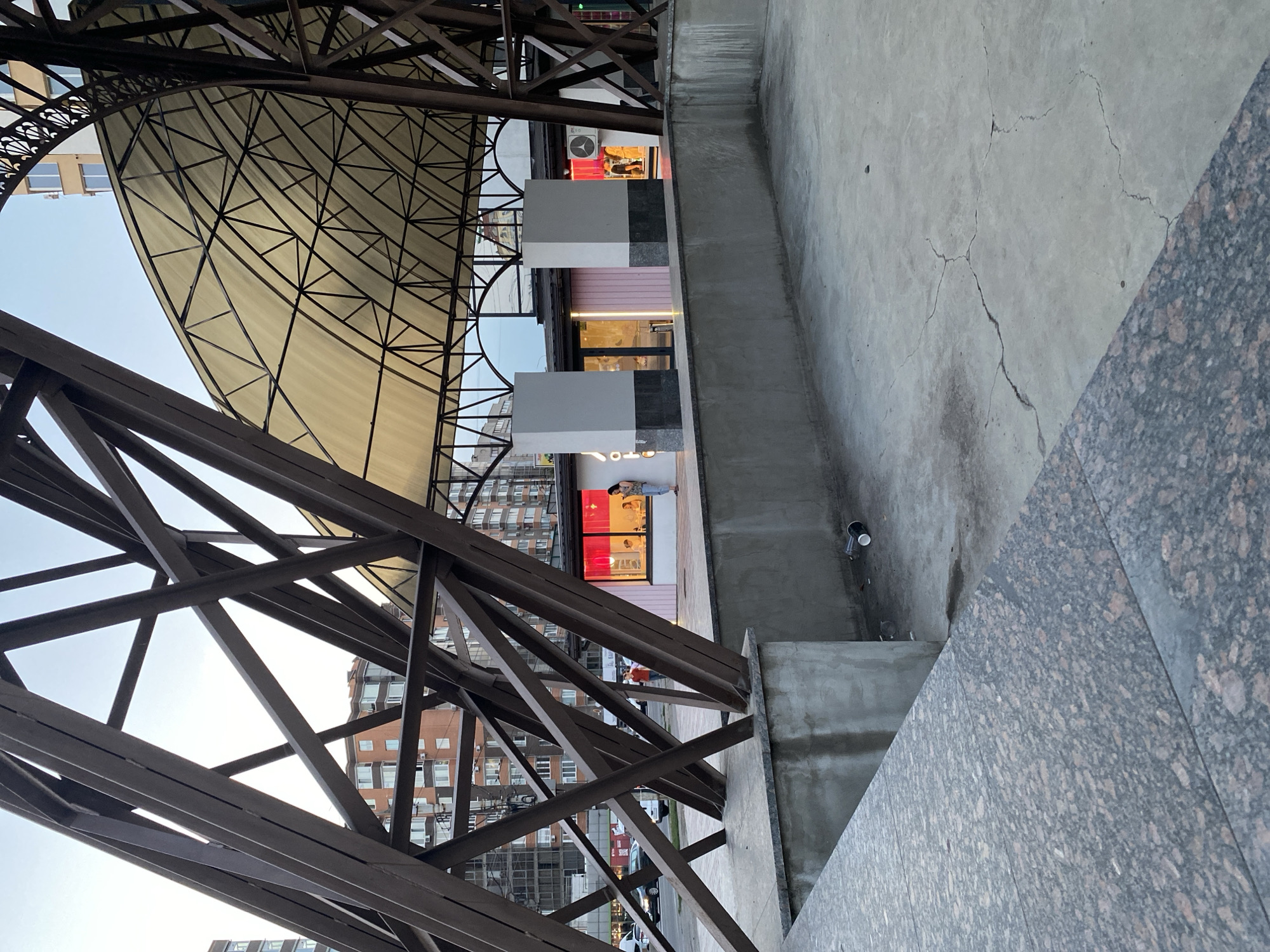 The image depicts a public space with architectural elements and urban surroundings in Vinnytsia, Ukraine. In the foreground, a textured granite surface leads to a concrete area with visible cracks and discarded paper cups. Behind this, a low concrete wall separates it from a paved area.
Dominating the mid-ground is a large, open-air structure with a metal, lattice-like framework supporting a translucent, light-colored canopy. Beneath the canopy, a row of businesses is visible. One establishment has large windows with bright pink and red lighting. Two figures are standing outside this establishment, and several other individuals are inside, illuminated by the interior lights.
Further back, a street with cars and buildings is visible. The buildings are multi-story structures, some with brick facades and numerous windows. The sky above is overcast, suggesting either early morning or late afternoon. The overall scene conveys a sense of urban infrastructure and commercial activity within Vinnytsia.
