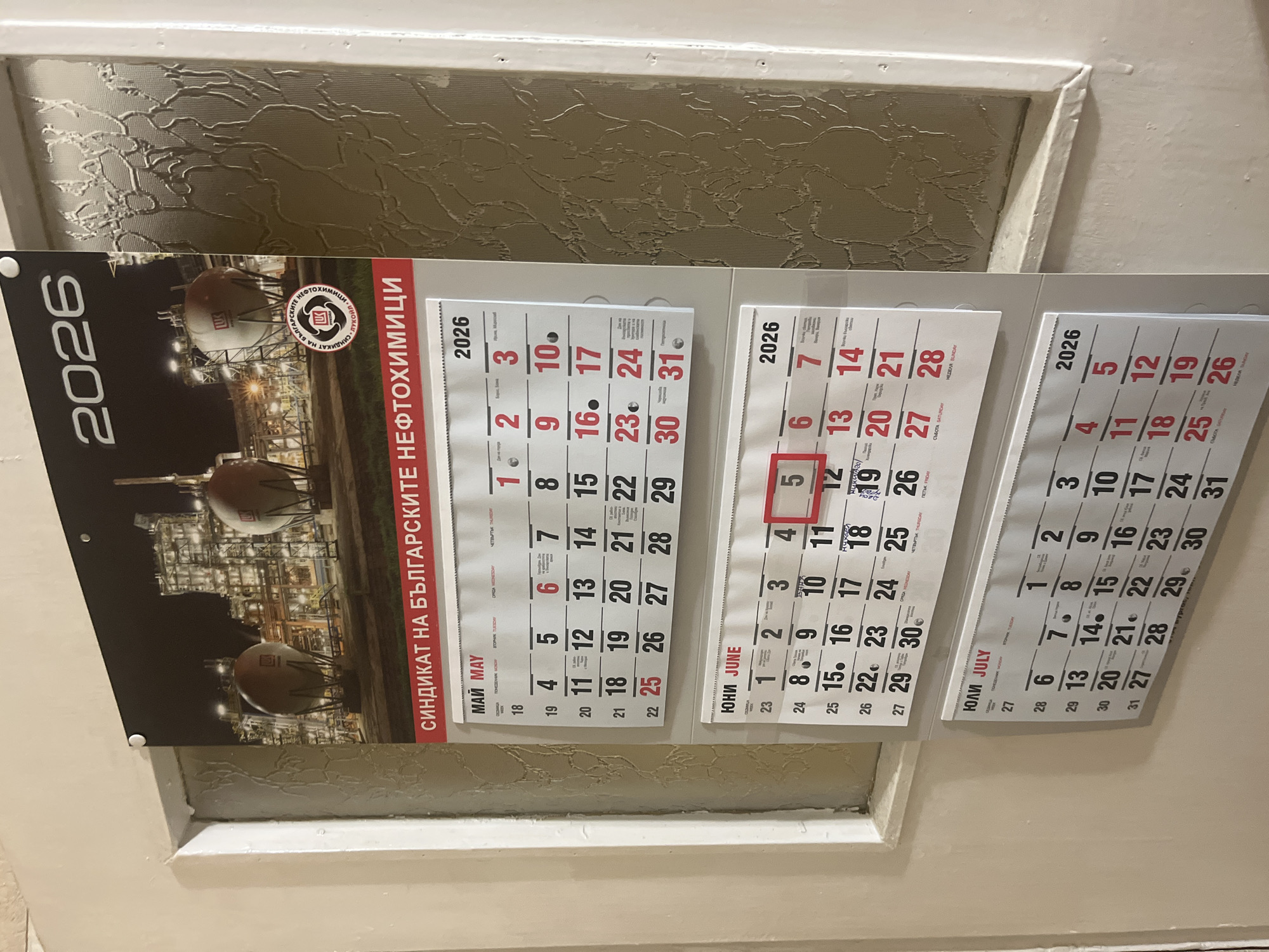 This is a full shot of a wall calendar hanging on a white wall next to a textured glass panel. The calendar features a photograph of an industrial facility at night, with large storage tanks illuminated. Text on the calendar is in Bulgarian, indicating it is from 2026. It has sections for May, June, and July. The calendar also displays the name "СИНДИКАТ НА БЪЛГАРСКИТЕ НЕФТОХИМИЦИ" which translates to "Union of Bulgarian Petrochemists". There is no information within the image to suggest a specific location in Burgas, Bulgaria.
