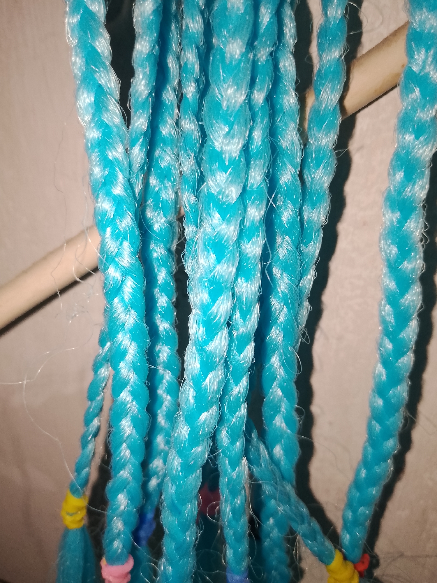 In Santa Ana, Venezuela, a vertical close-up shot captures multiple strands of synthetic, bright turquoise braided material, each composed of a three-strand plait. These braids are suspended from a horizontal, light-colored wooden rod visible in the upper portion of the frame. The synthetic fibers exhibit a distinct sheen, and minor wisps of loose material are discernible along the braids. Towards the lower ends of several braids, small, vibrant embellishments are present, including yellow elastic bands and pink spherical beads. The background consists of a lightly textured, out-of-focus beige or light grey wall, with soft shadows cast upon it from an unseen light source.