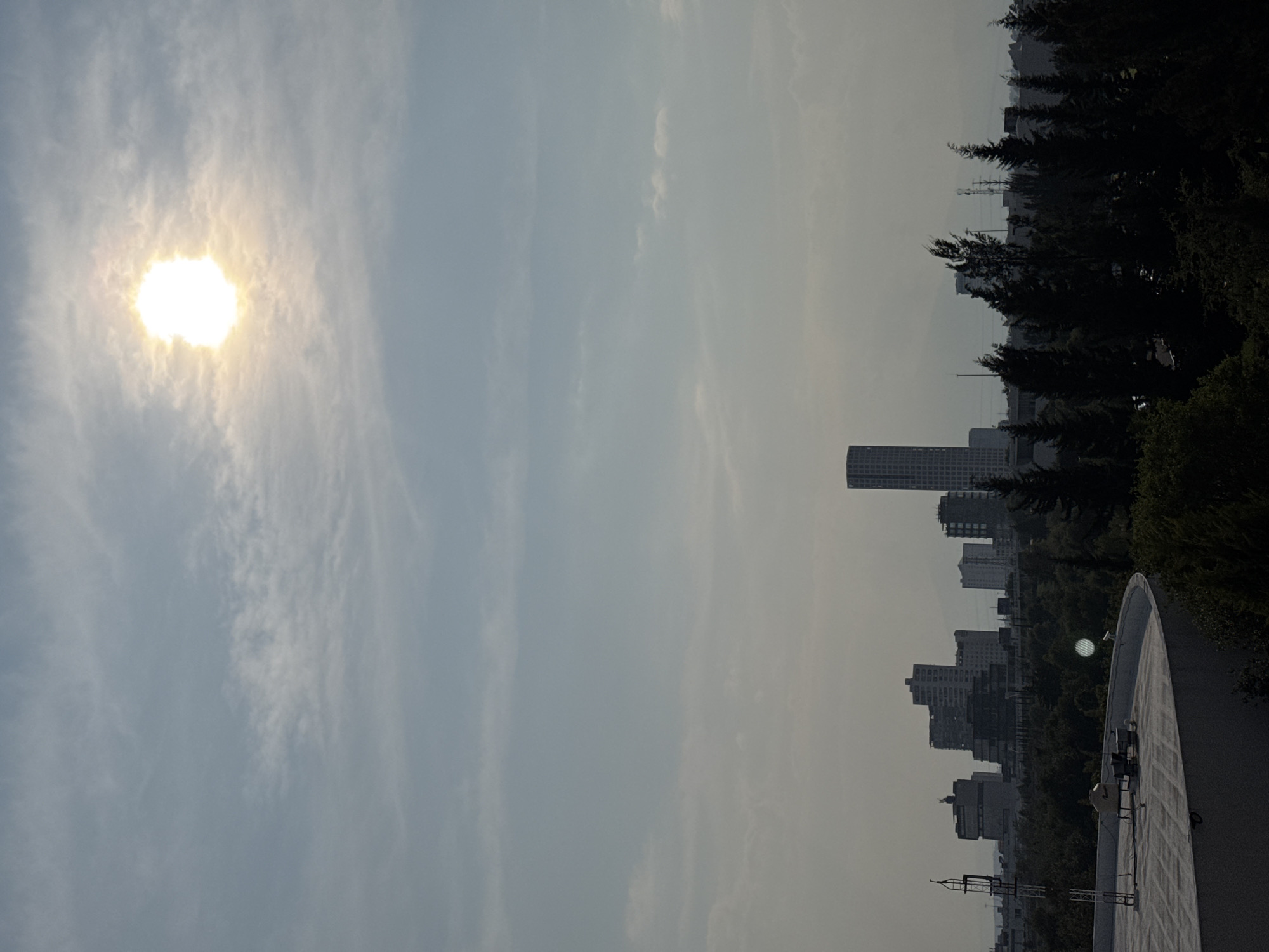 The image shows a view from a rooftop in the city of Heroica Puebla de Zaragoza, Mexico. The sky is predominantly a hazy blue with thin, scattered clouds. The sun is high in the sky, positioned on the left side of the frame. In the background, a cityscape is visible, including several high-rise buildings and trees. In the foreground is a portion of a rooftop with visible structures.