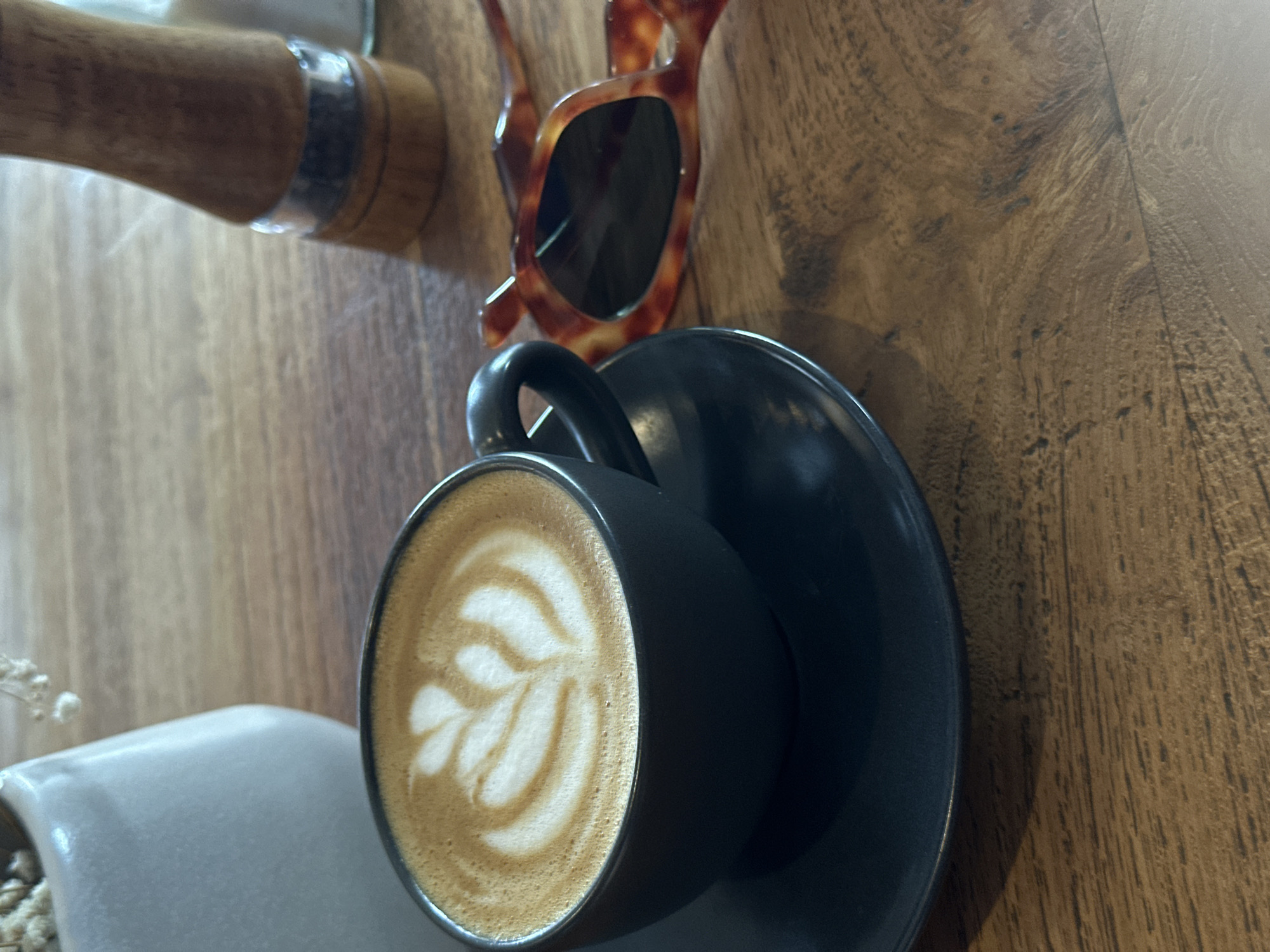 The image captures a coffee cup with latte art, resting on a black saucer on a wooden table. Beside it are sunglasses, part of a tamper, and what appears to be a small vase with dried flowers. The scene is likely in North Kuta, Indonesia. There are no people visible.