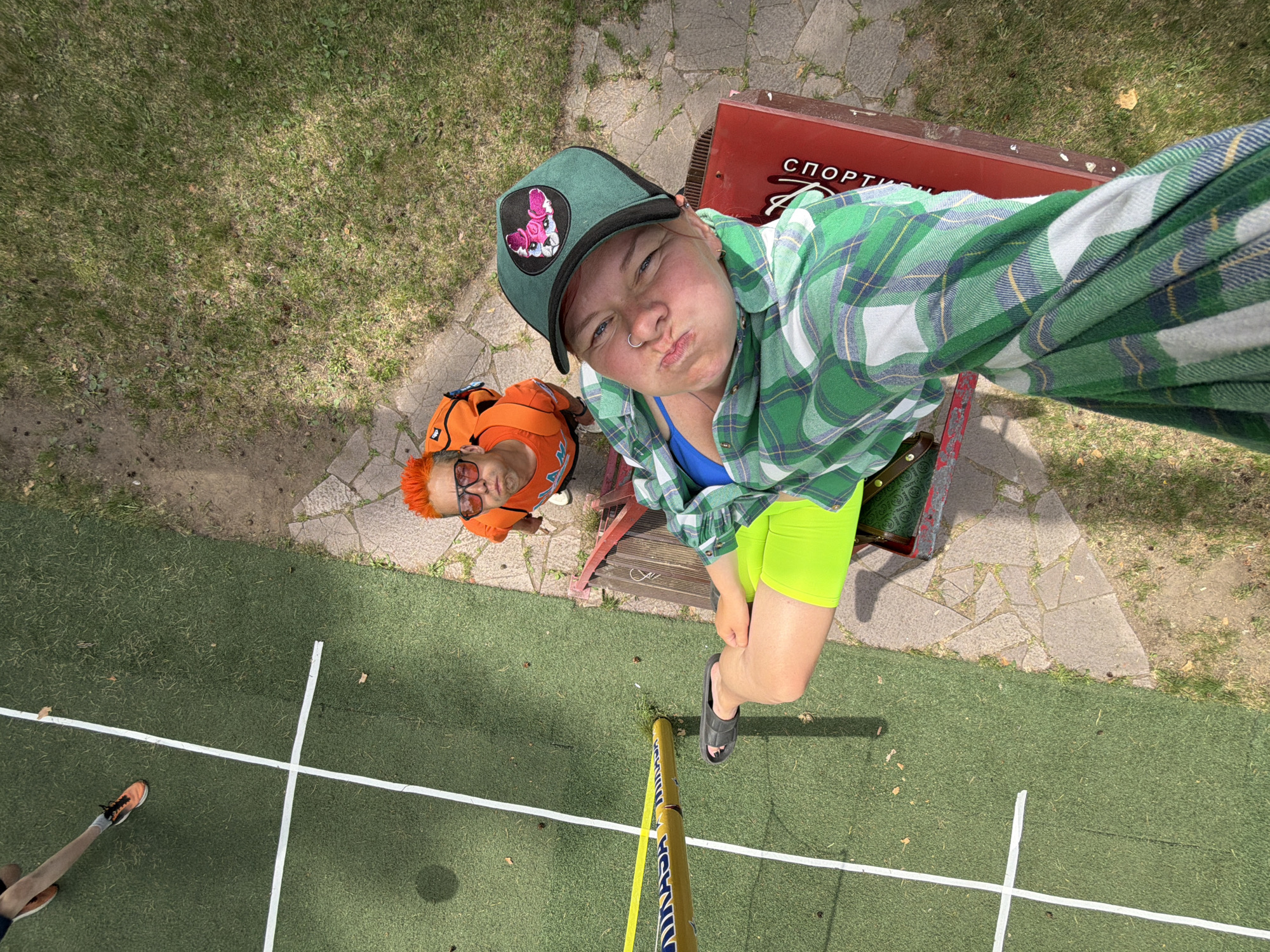 A high-angle photograph depicts two individuals next to a low red structure with Cyrillic text, specifically the word "СПОРТ" ("SPORT" in English), potentially indicating its function or purpose. The photograph was taken in the city of Chaykovsky, Russia.
One person is taking a selfie. She wears a green cap with a patch and a green and white flannel shirt over a blue top. She is wearing neon green shorts and dark sandals. Her expression is pouting.
Another person, likely on the ground, is clad in an orange top and has orange hair, and wears sunglasses.
The setting appears to be an outdoor sports area. The ground is covered with green artificial turf. There are painted white lines marking a playing field. Part of a yellow pole lies in front of the first person. The edge of a walkway made of stone slabs is visible.